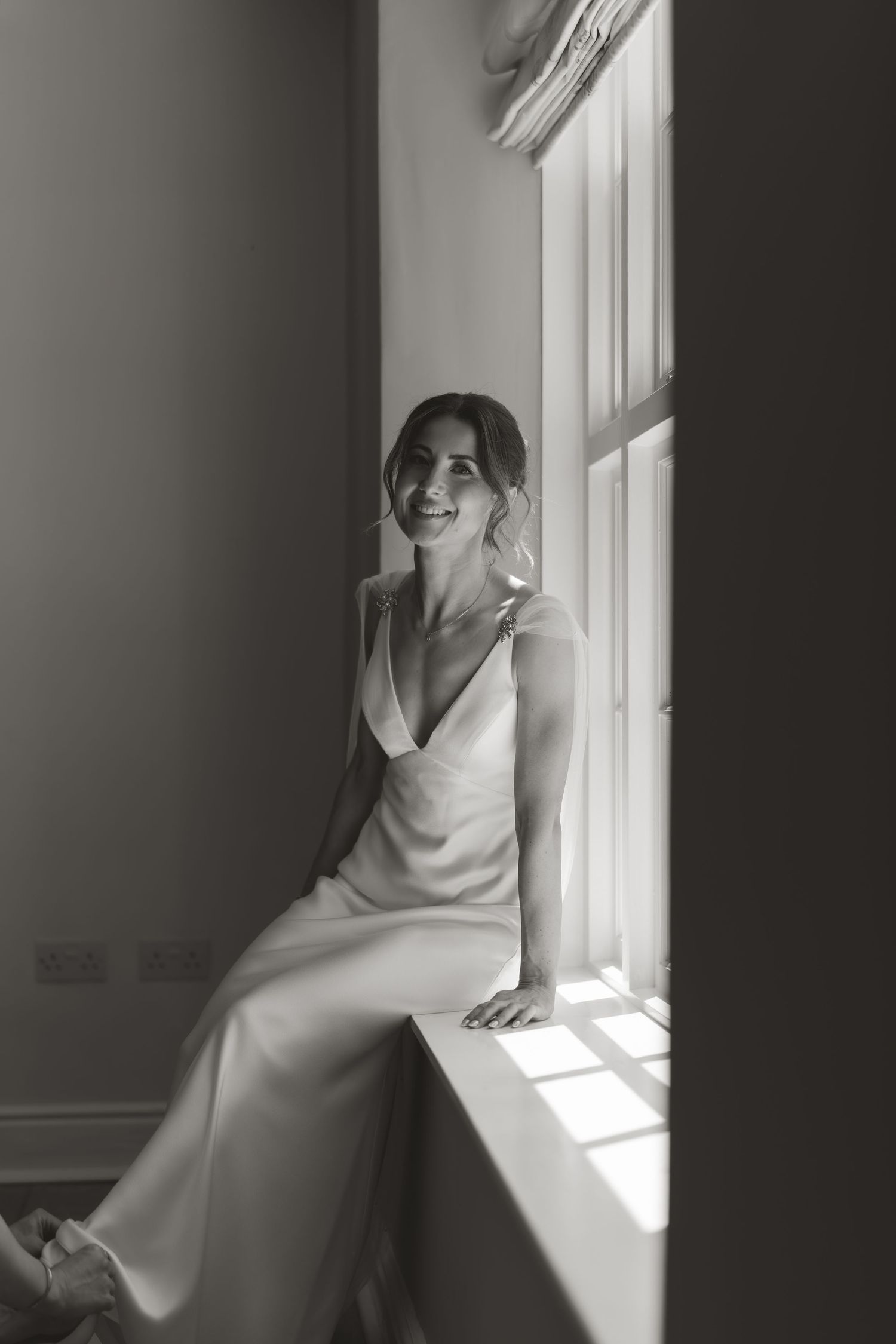 Black and white portrait of bride sitting on windowsill in flowing white gown with natural light streaming in.
