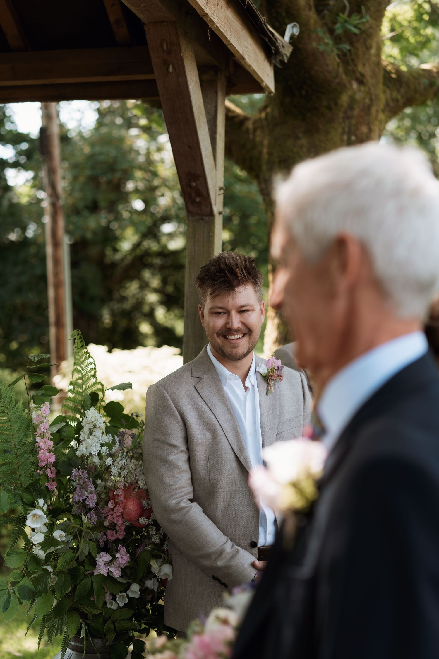 Wedding ceremony moment with guests and floral arrangements under a rustic wooden structure.