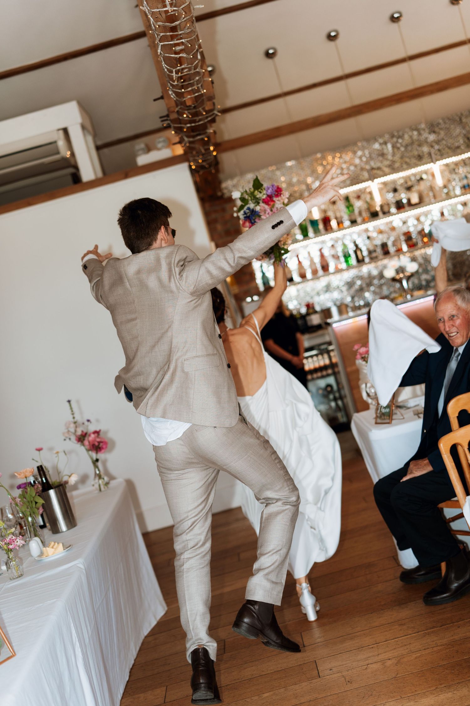 Wedding guests dancing in white attire on a wooden floor at an indoor reception venue.