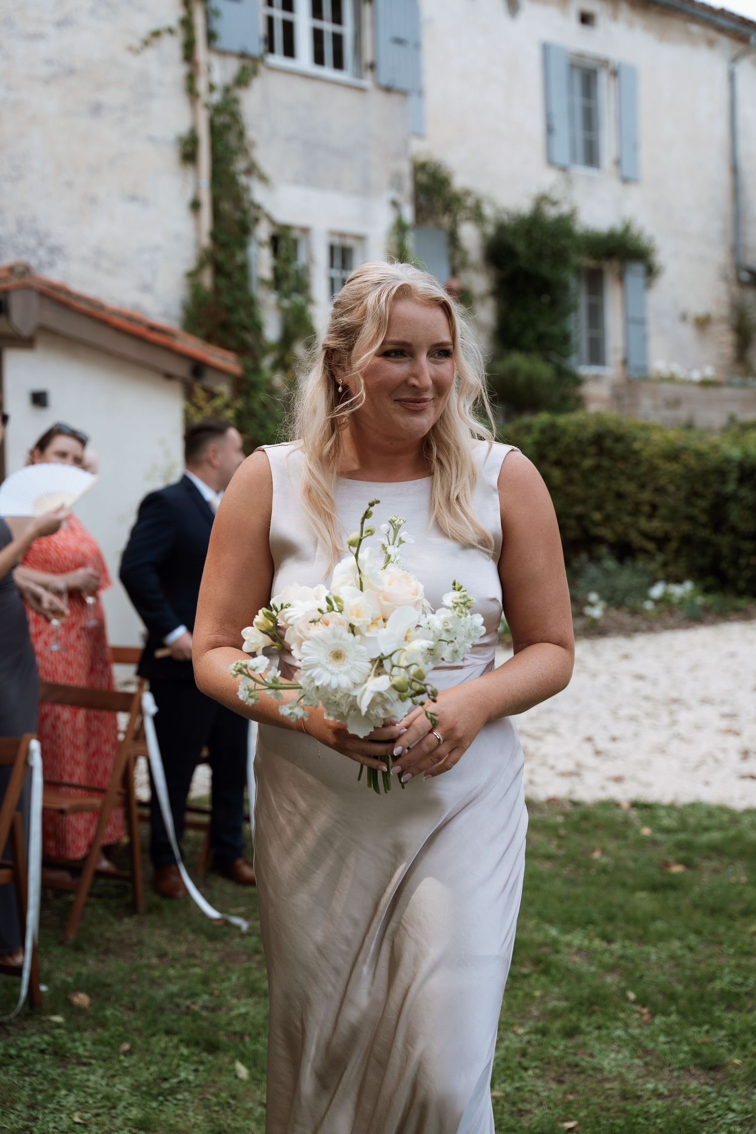Person in white dress holding delicate white floral bouquet against backdrop of rustic countryside building.
