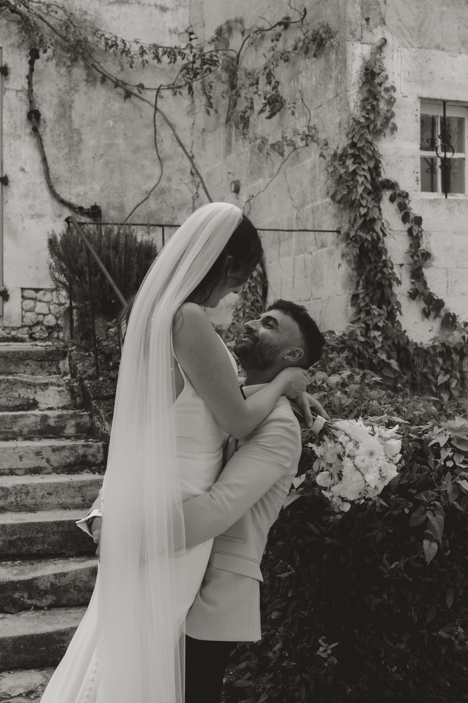 Black and white photo of couple embracing during wedding celebration against stone wall with vines.