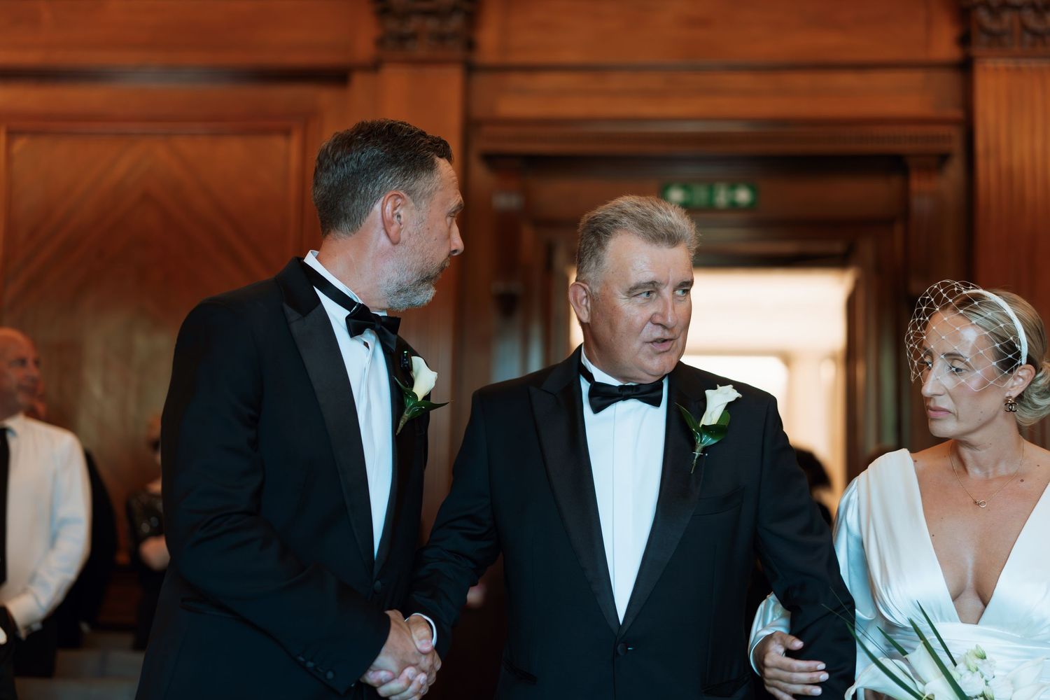 Three people in formal black tie attire engaged in conversation at an elegant indoor venue.