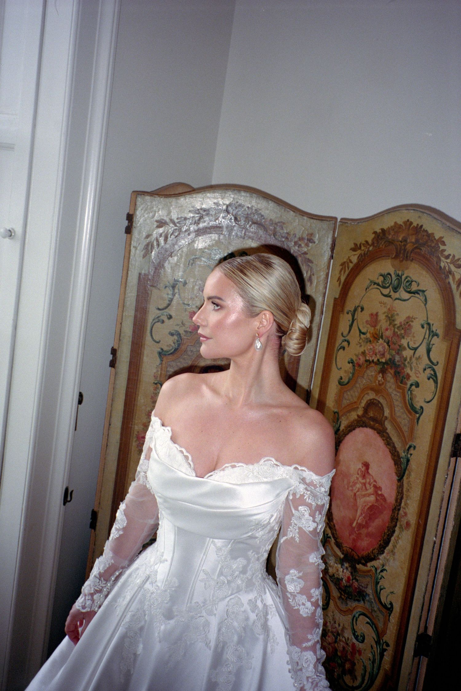 A wedding dress with delicate lace sleeves and off-shoulder neckline shown in multiple angles against a decorative screen.
