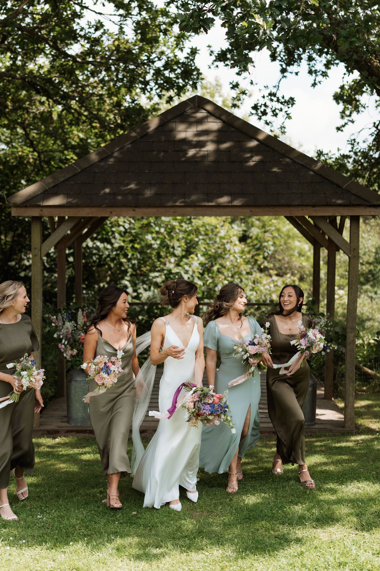 Wedding party relaxing and laughing together under a wooden pavilion on a sunny afternoon.