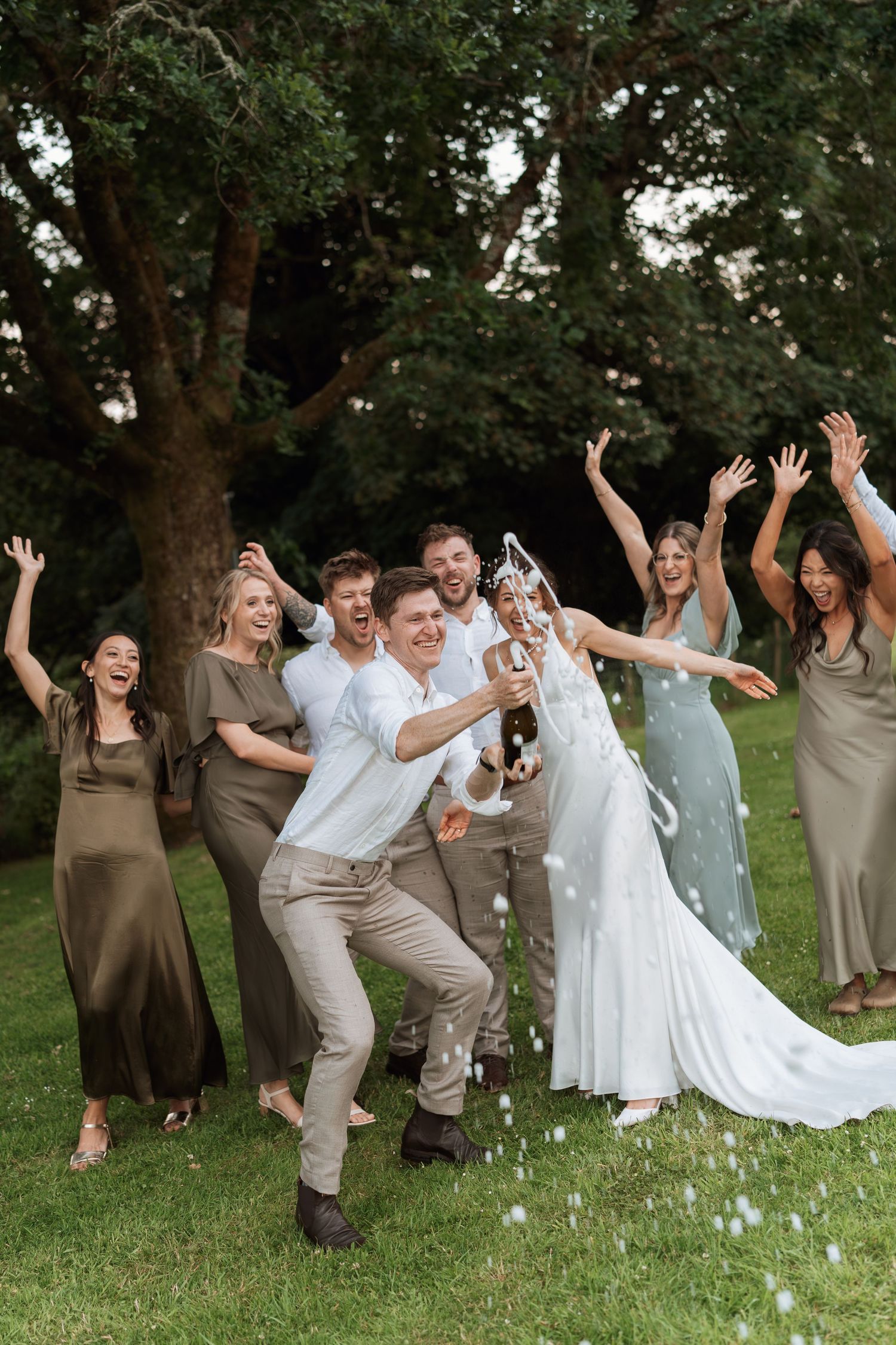 Wedding party celebrates joyfully on grass with arms raised wearing neutral toned attire.