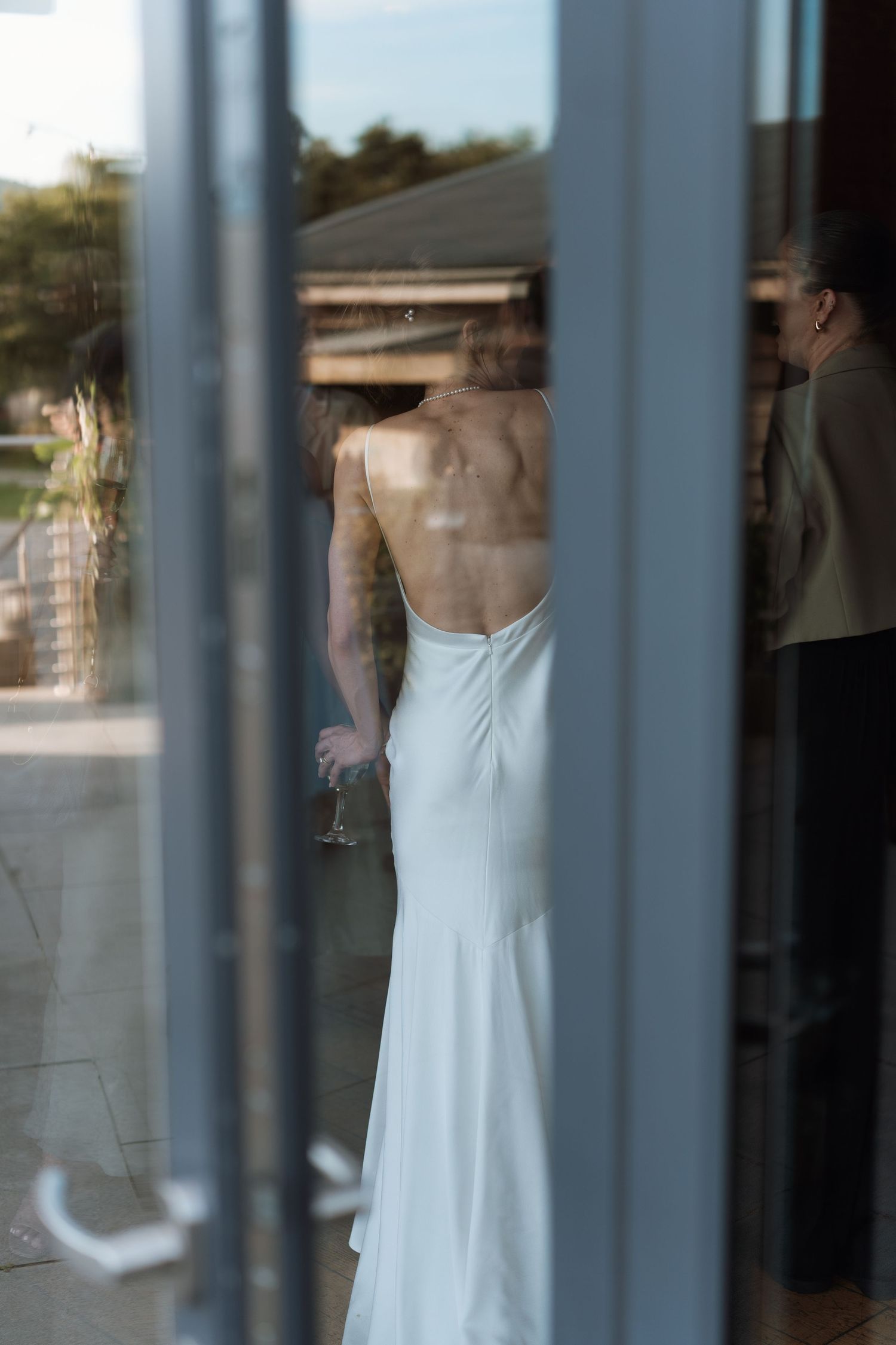 Through window panes, a figure in a white formal dress is seen from behind during an elegant indoor gathering.