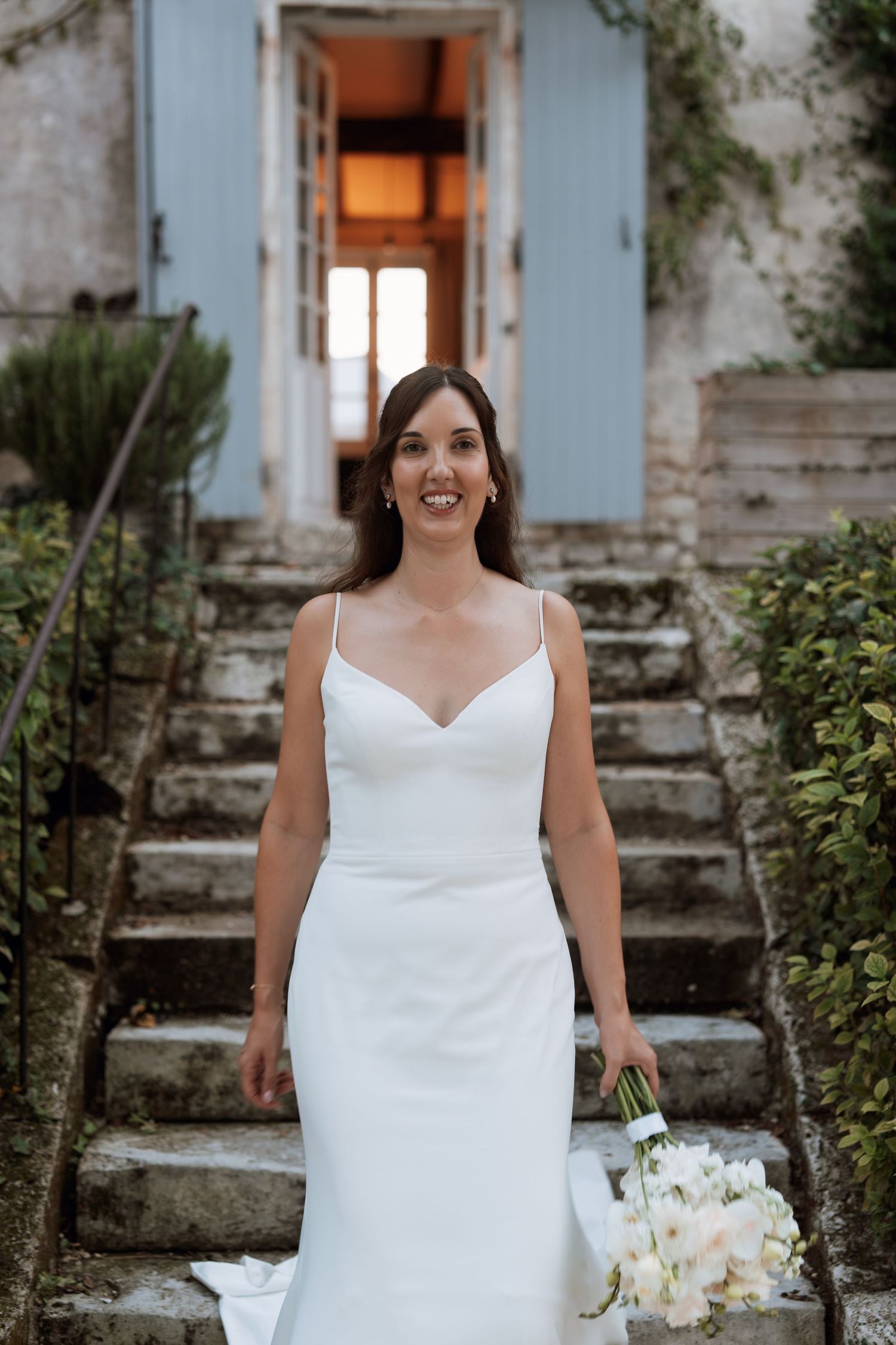 A bride in a sleek white wedding dress holds a bouquet of white flowers on stone steps.