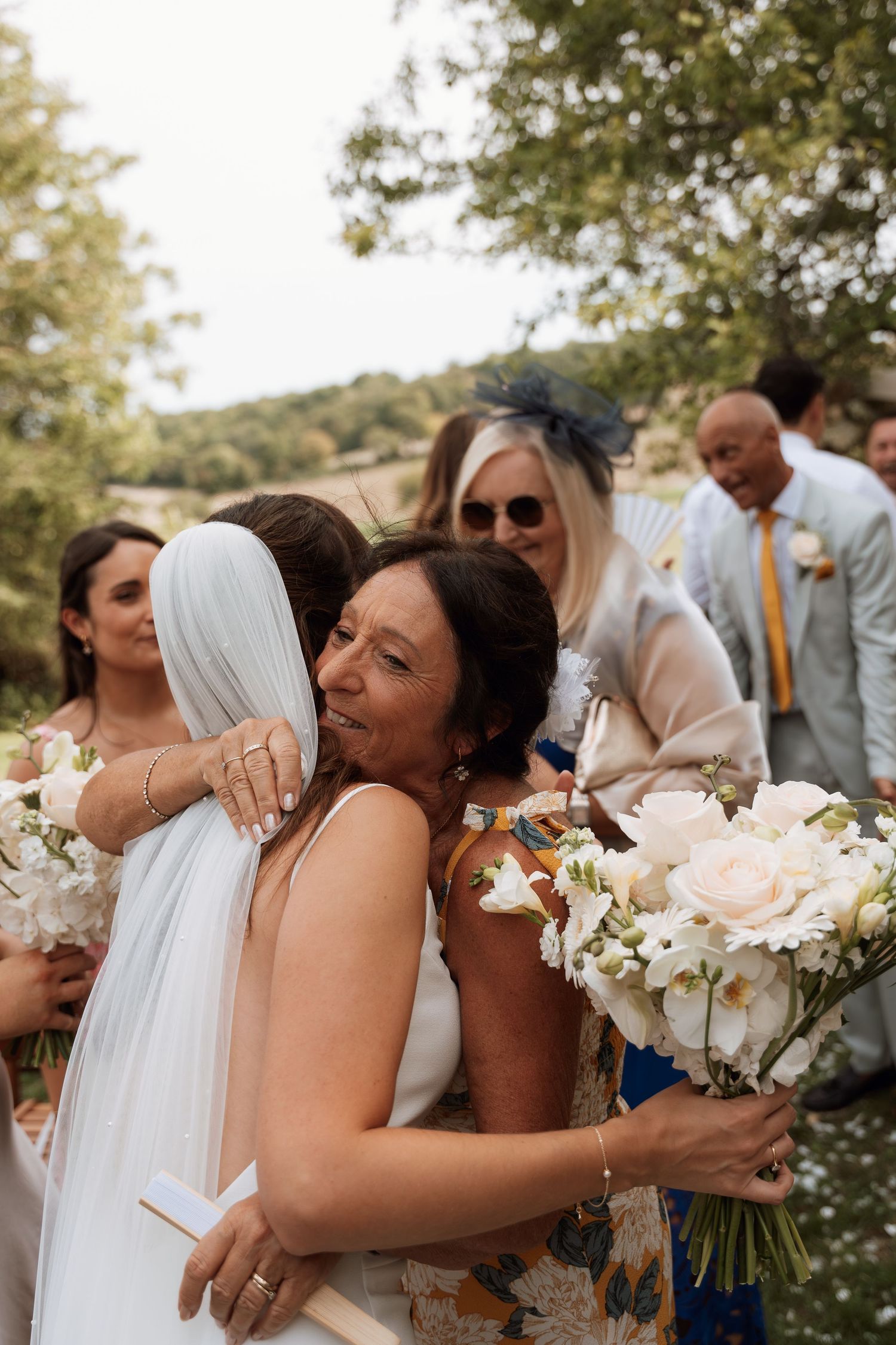 Emotional embrace between bride and wedding guests with white floral bouquets outdoors.