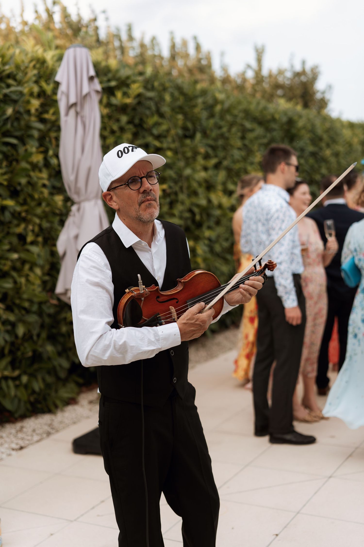 Musician in white shirt and black vest plays violin during outdoor wedding reception.