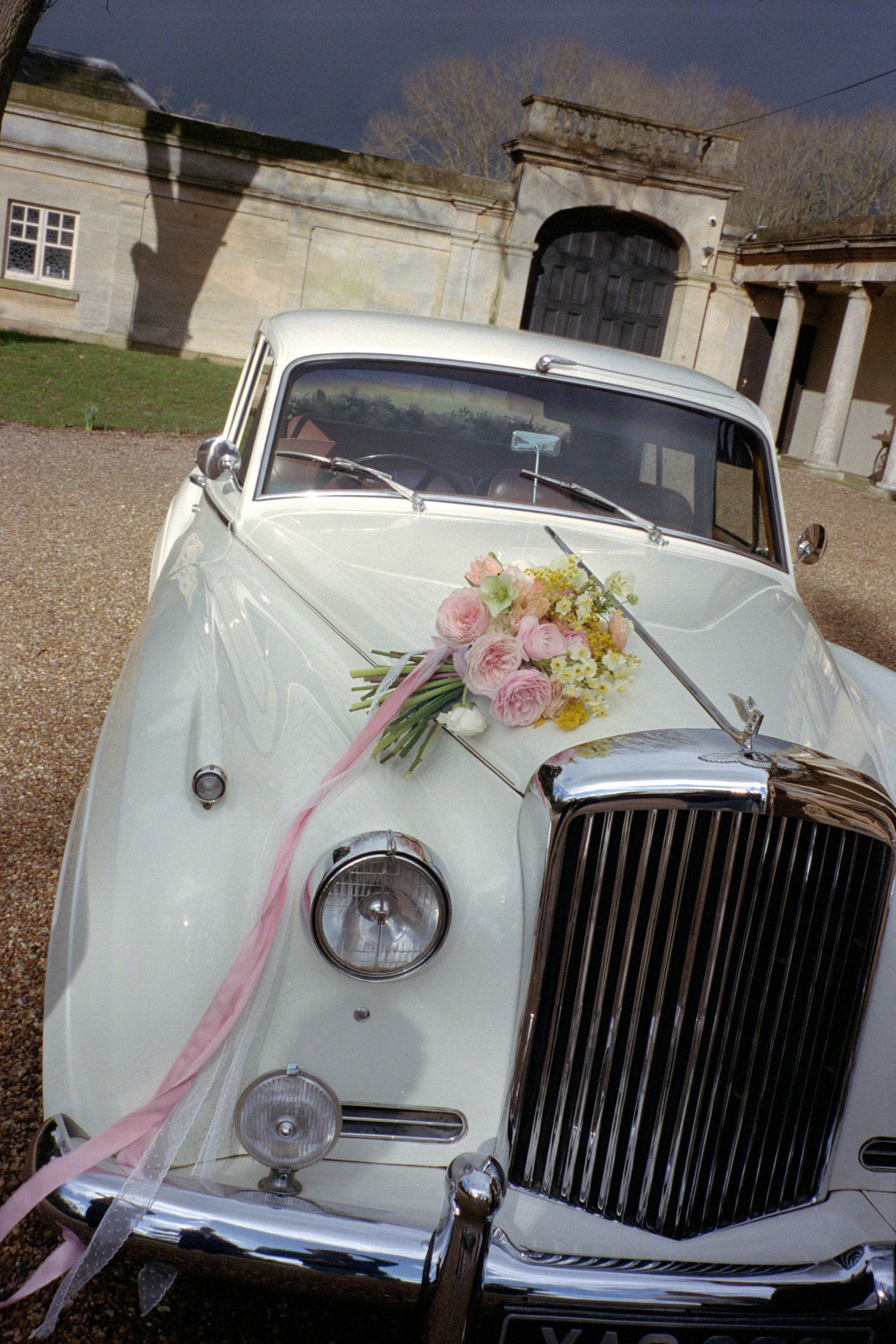 White vintage Rolls Royce wedding car decorated with pink and yellow floral arrangements and ribbons.