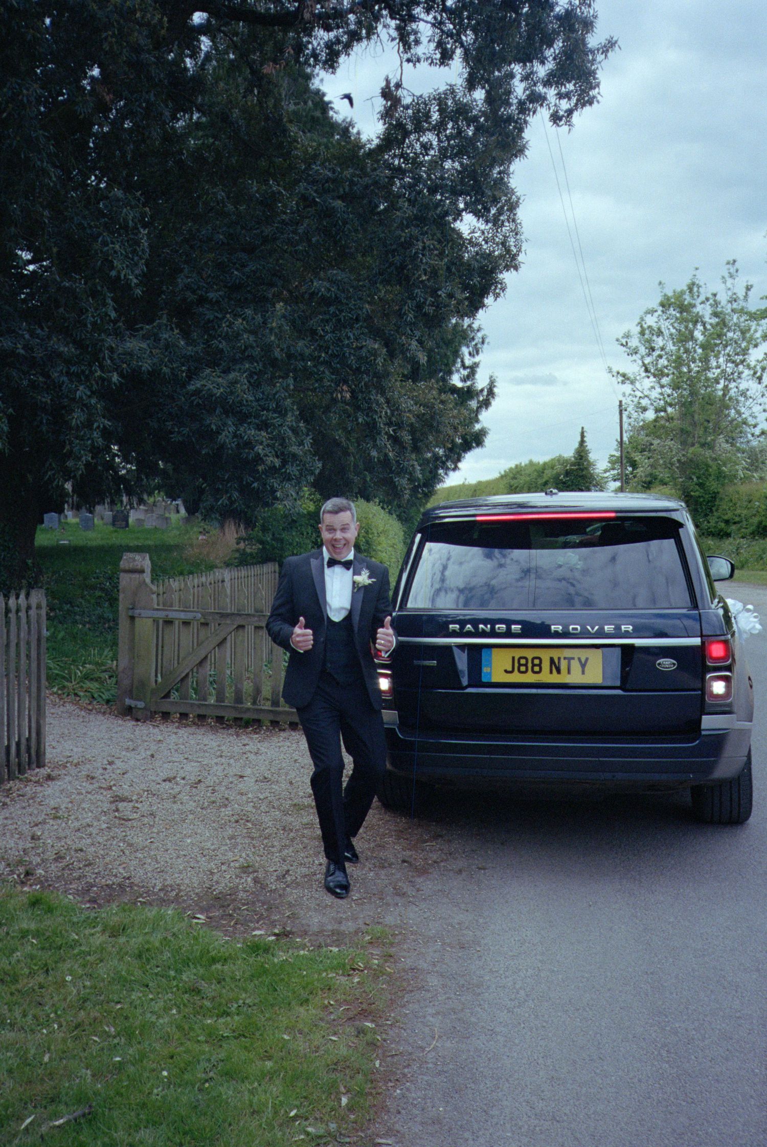 A person in a tuxedo walks past a black Range Rover on a rural path near a wooden fence.