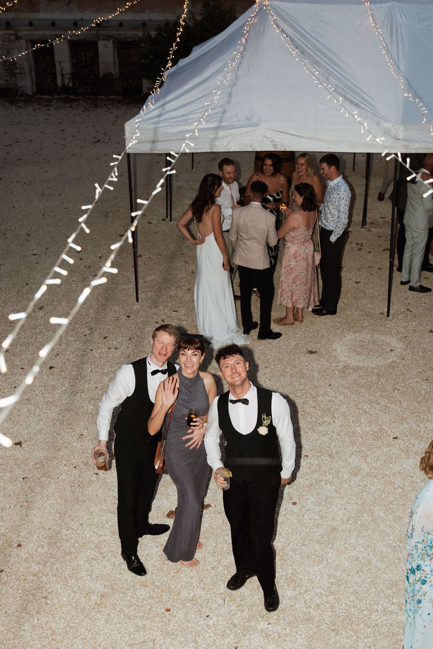 Wedding reception scene under a white tent with string lights and guests mingling.