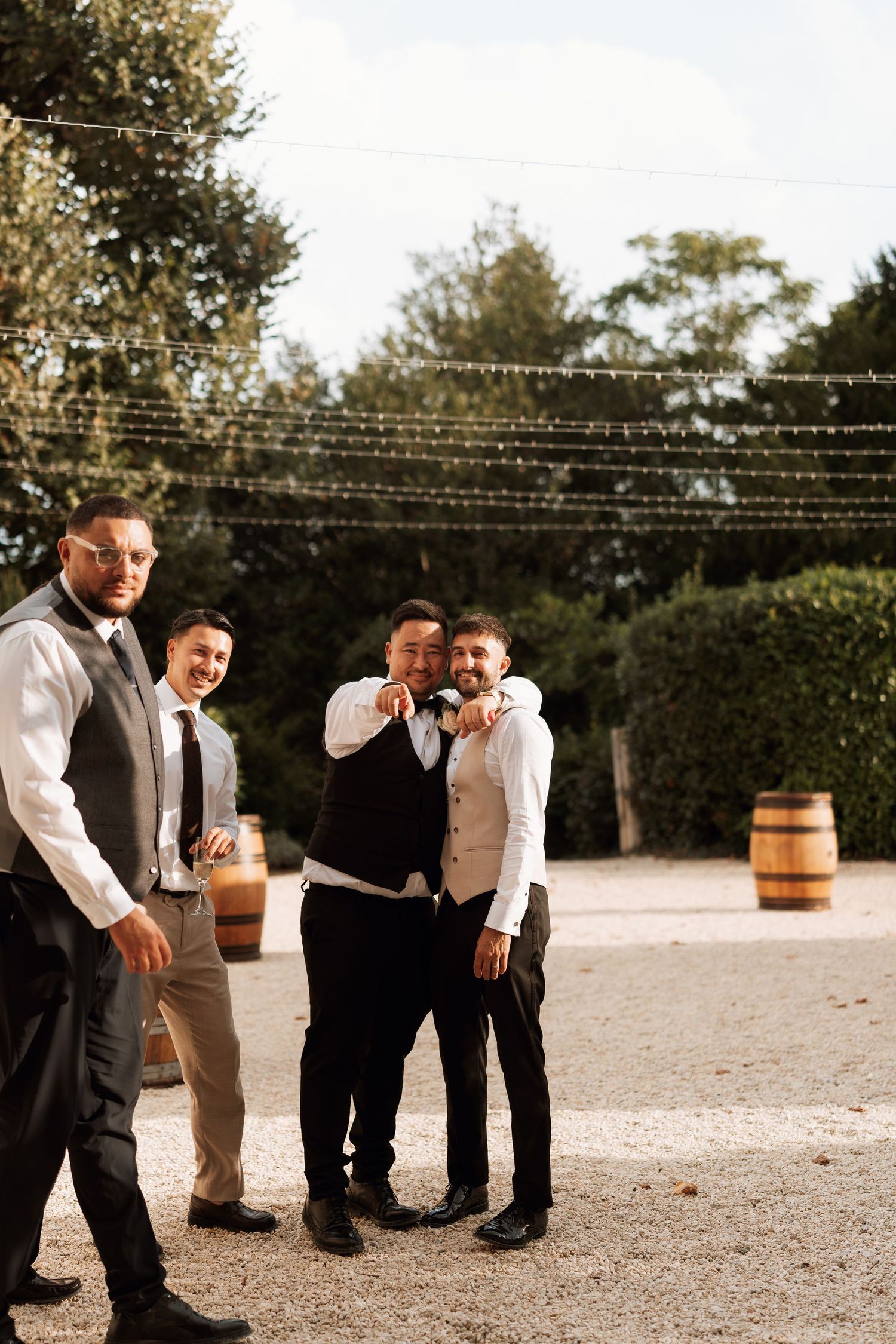 Groomsmen in formal attire gather outdoors under string lights at sunset.