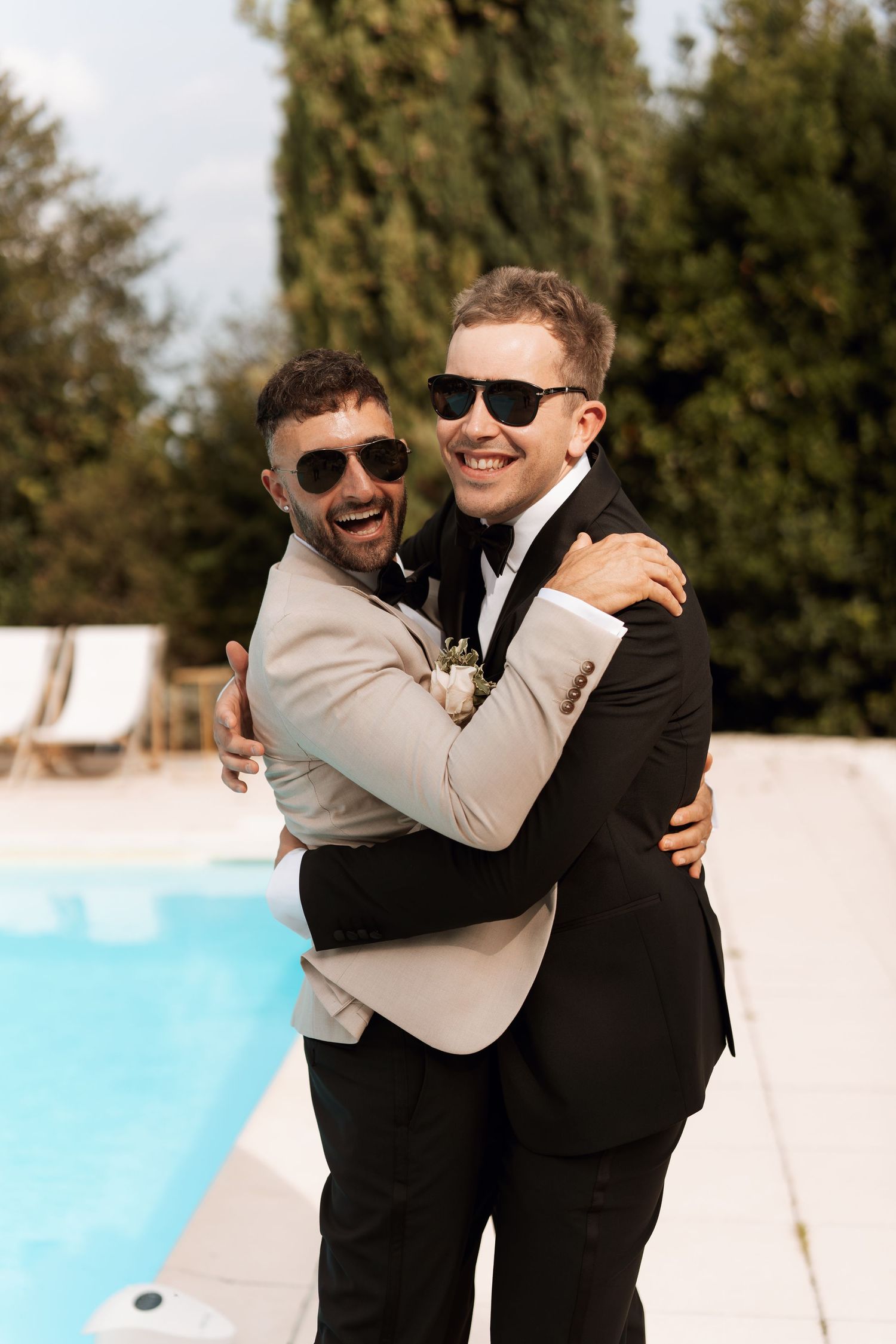 Two people in formal attire embrace and laugh by a pool.