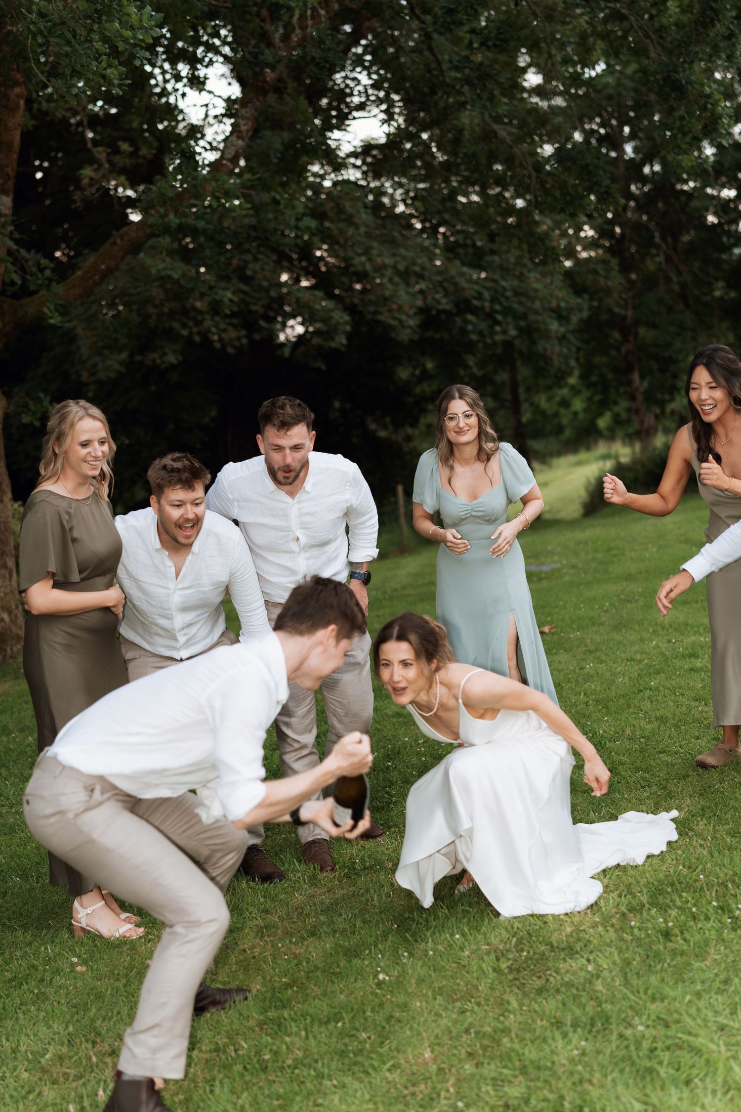 Wedding party performs energetic dance moves on grass during outdoor celebration.