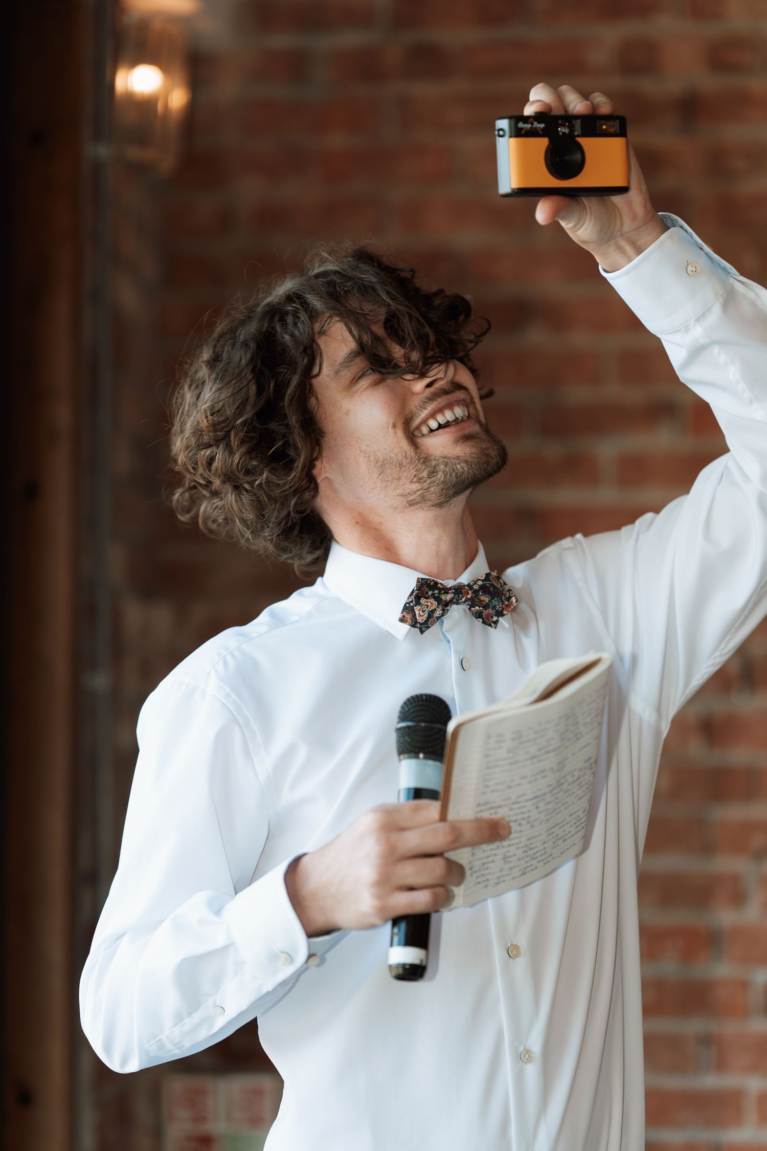Person in white shirt and bowtie performing energetically with microphone against brick wall.