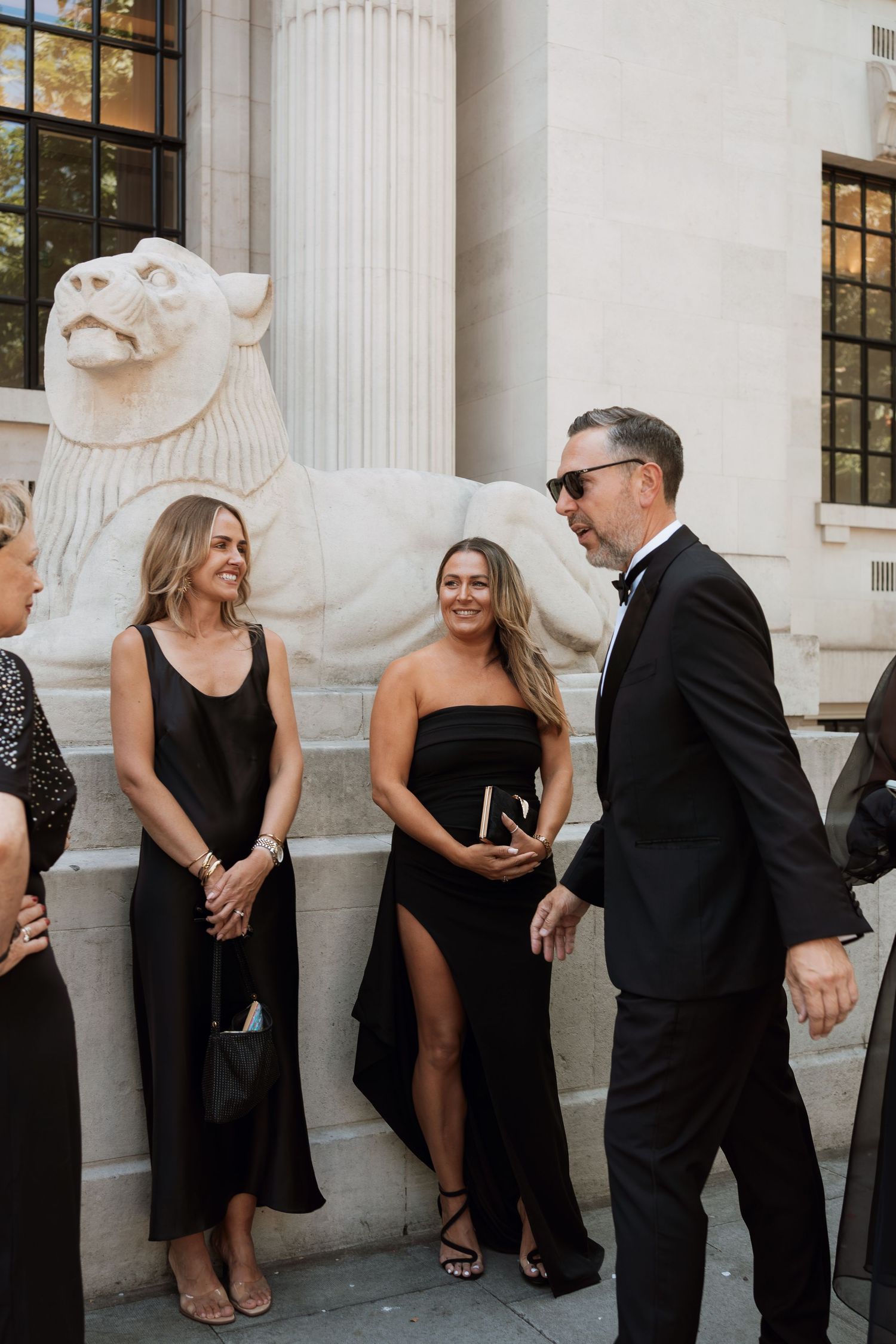 A group in formal attire stands chatting and laughing next to a decorative stone lion statue outside a building.