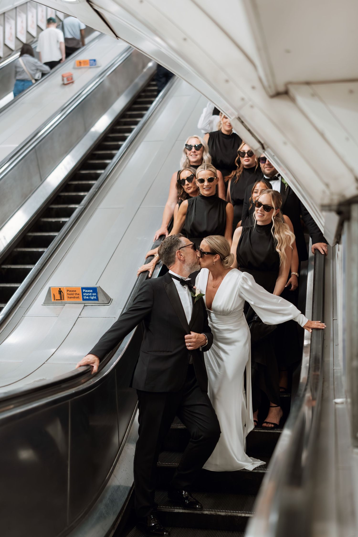 Wedding party in black and white attire poses on an escalator for a stylish photo.