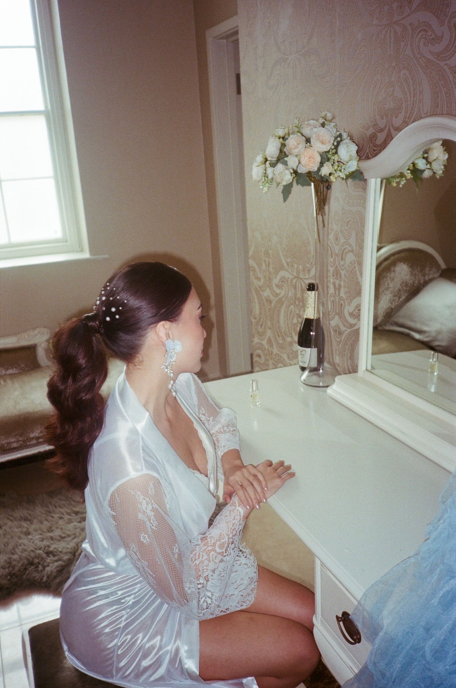 A wedding preparation scene in a white room with floral arrangements and natural light streaming through windows.