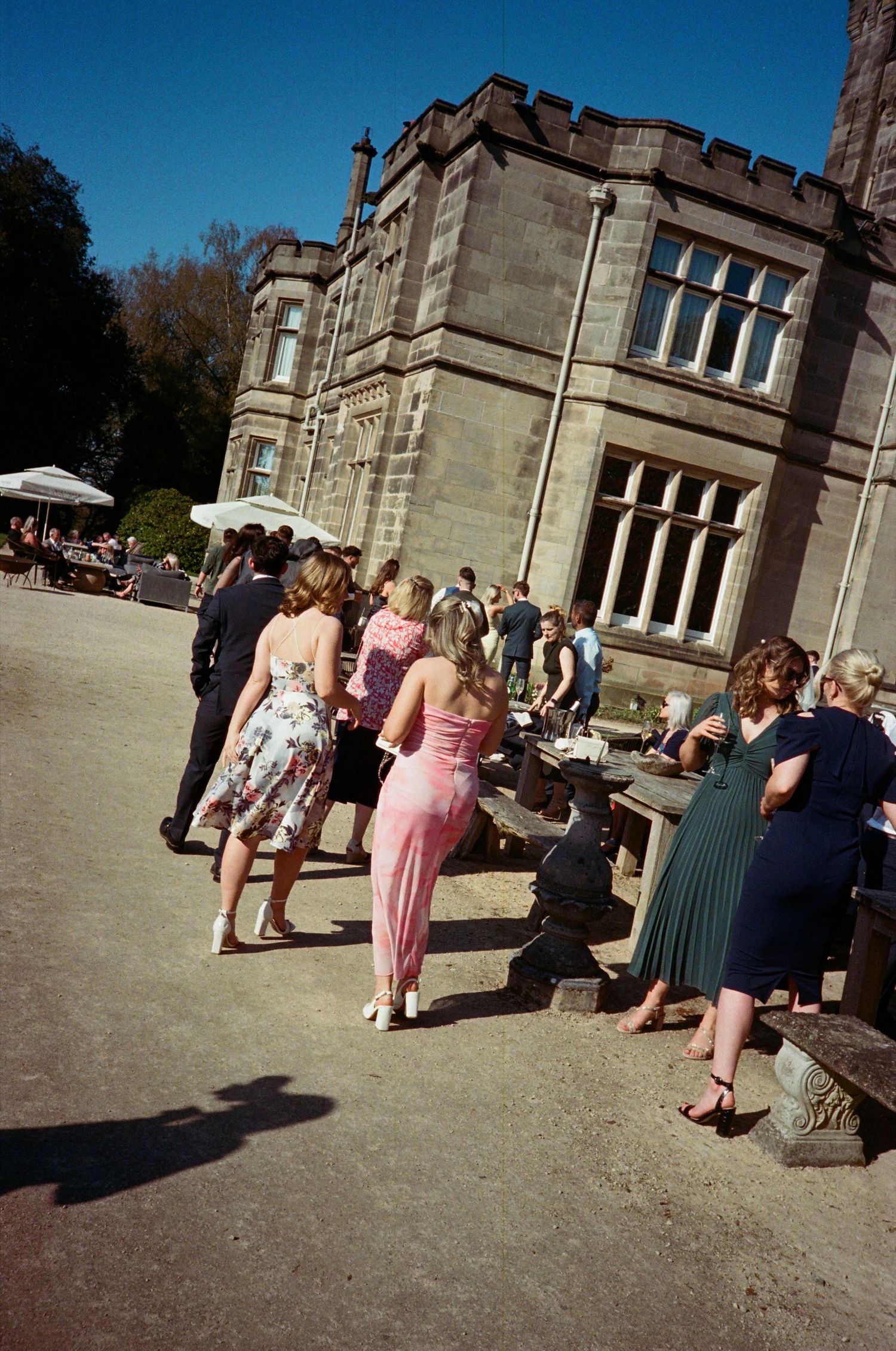 Guests gather outside a grand stone mansion for an elegant outdoor event on a sunny day.