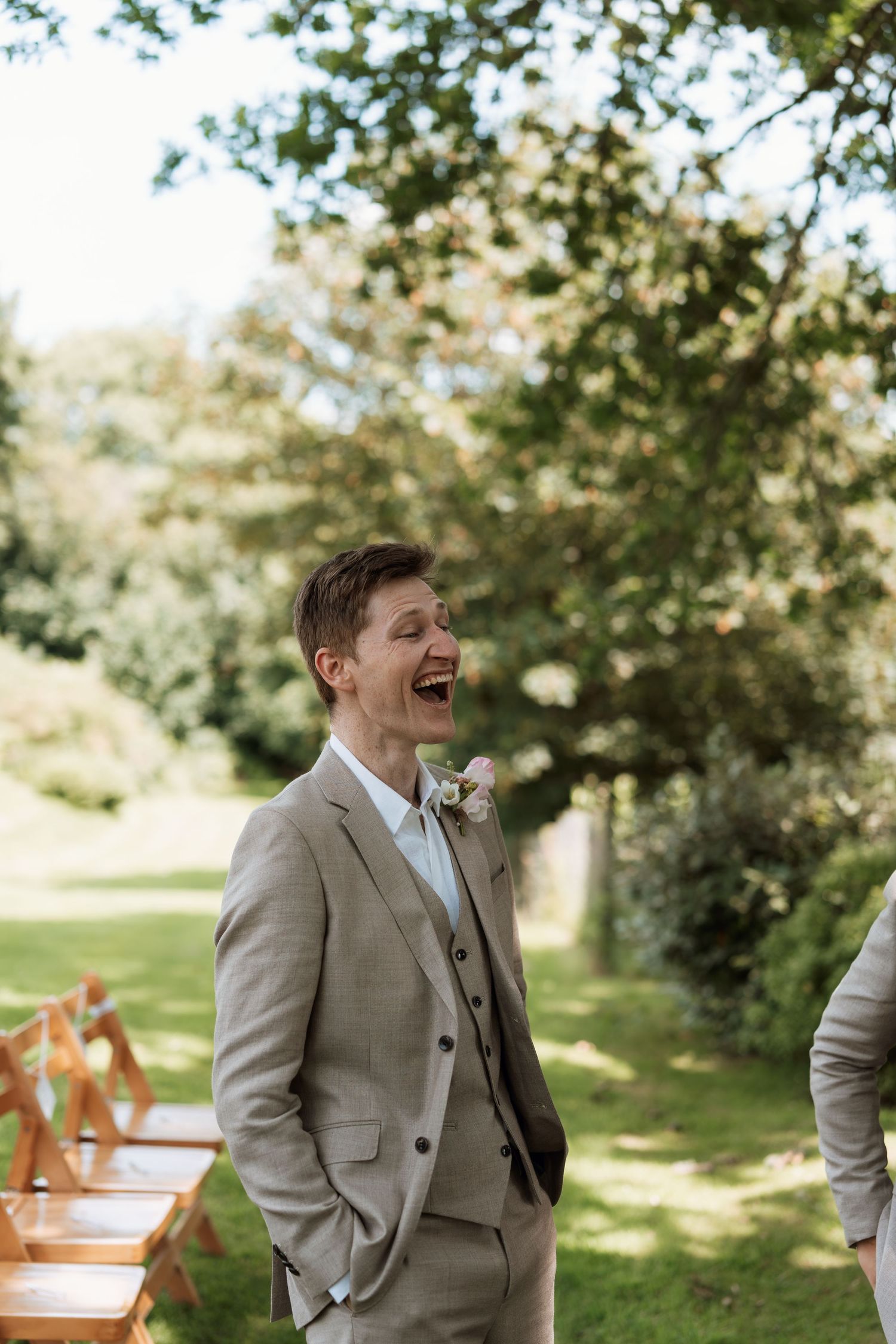 Person in light gray suit laughing joyfully in an outdoor garden setting.