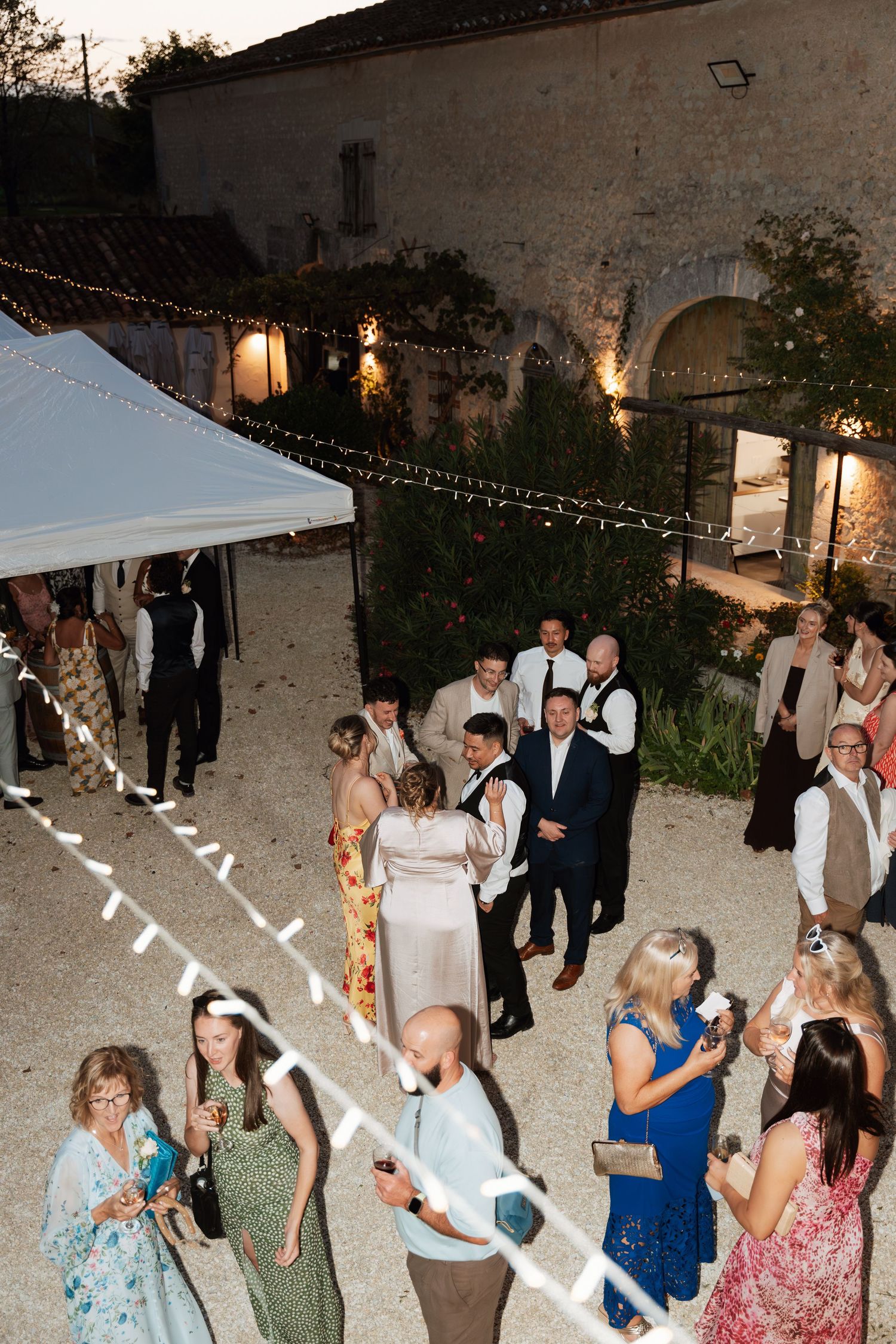 Wedding guests dancing outdoors at night under string lights near a stone building.