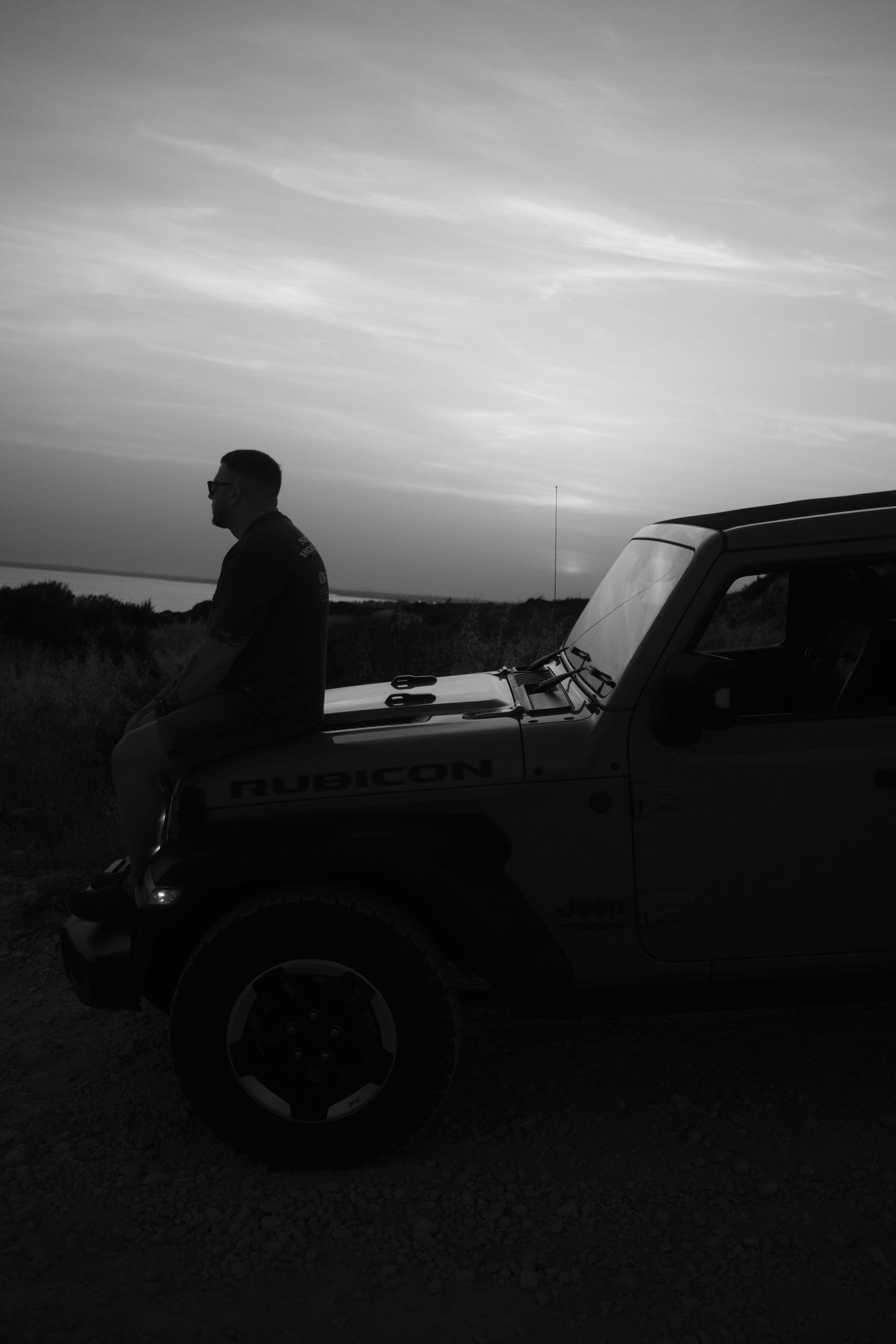 Silhouette of a man sitting on the hood of a Jeep Wrangler Rubicon at sunset in black and white.