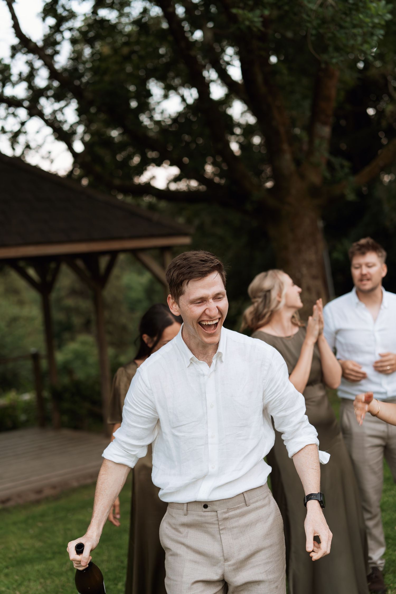 Person in white shirt and khaki pants laughs joyfully during outdoor wedding celebration.