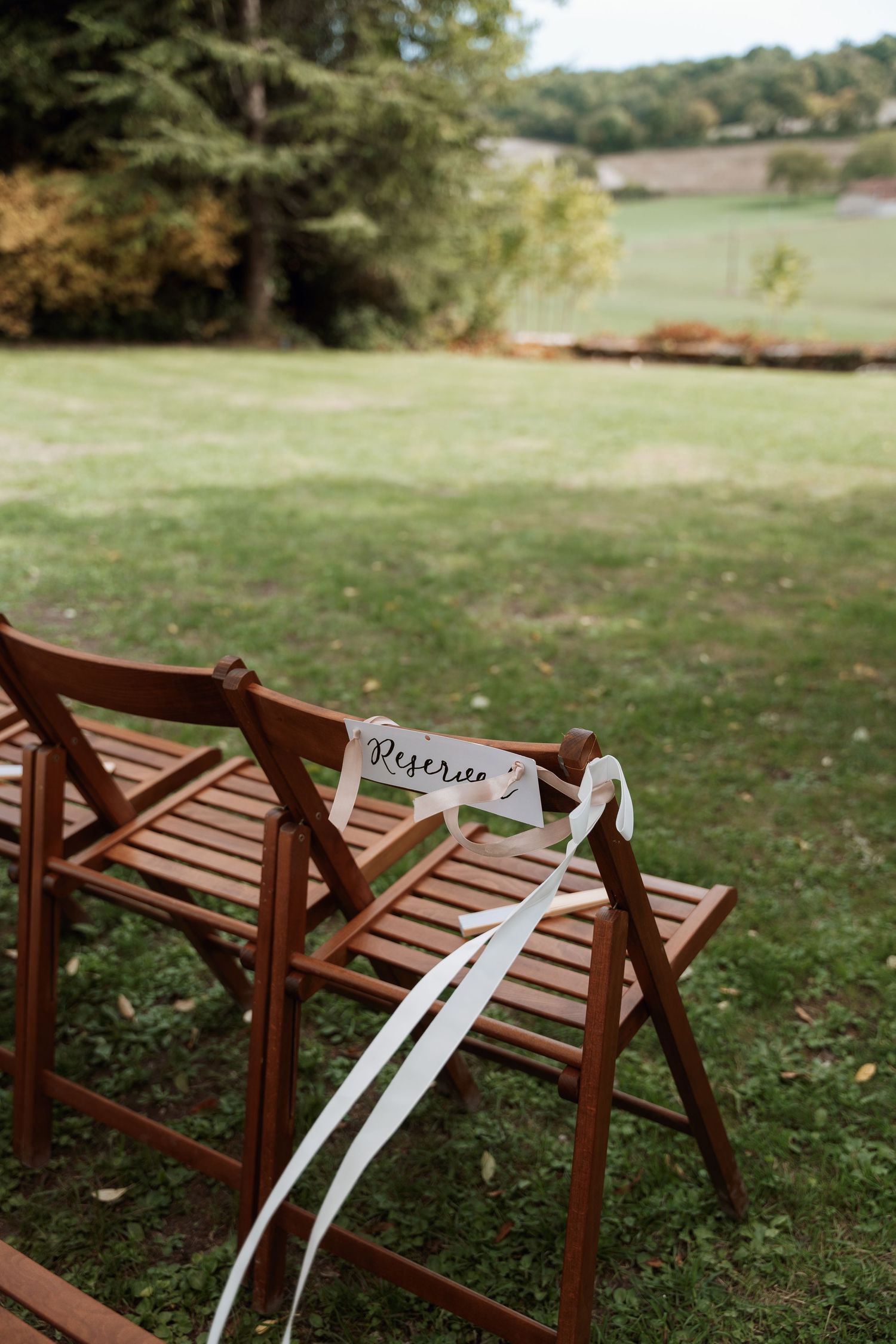 Wooden folding chairs with white ribbons set up on grass for outdoor ceremony.
