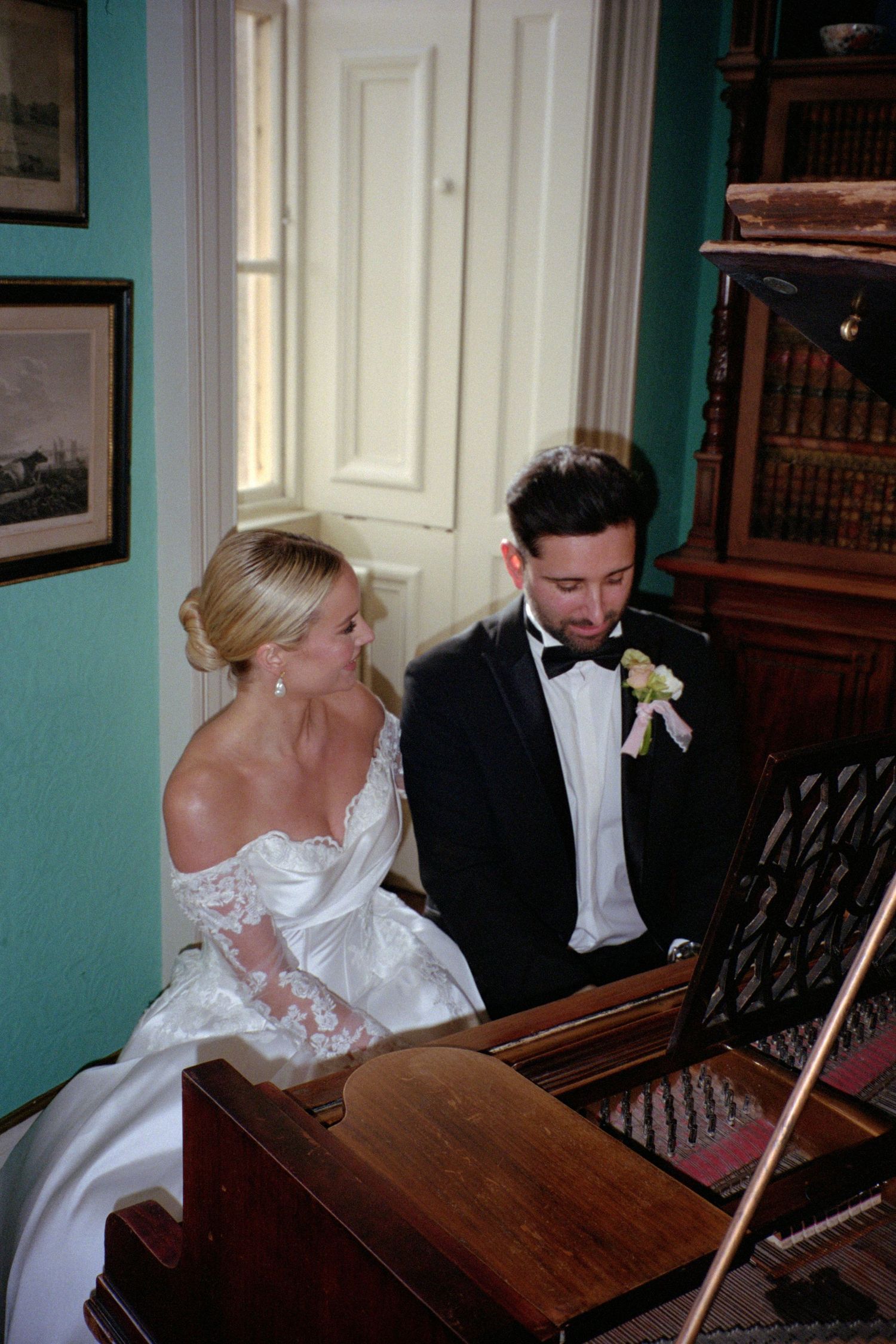 A bride and groom in formal attire sit together at a wooden piano in a room with turquoise walls.