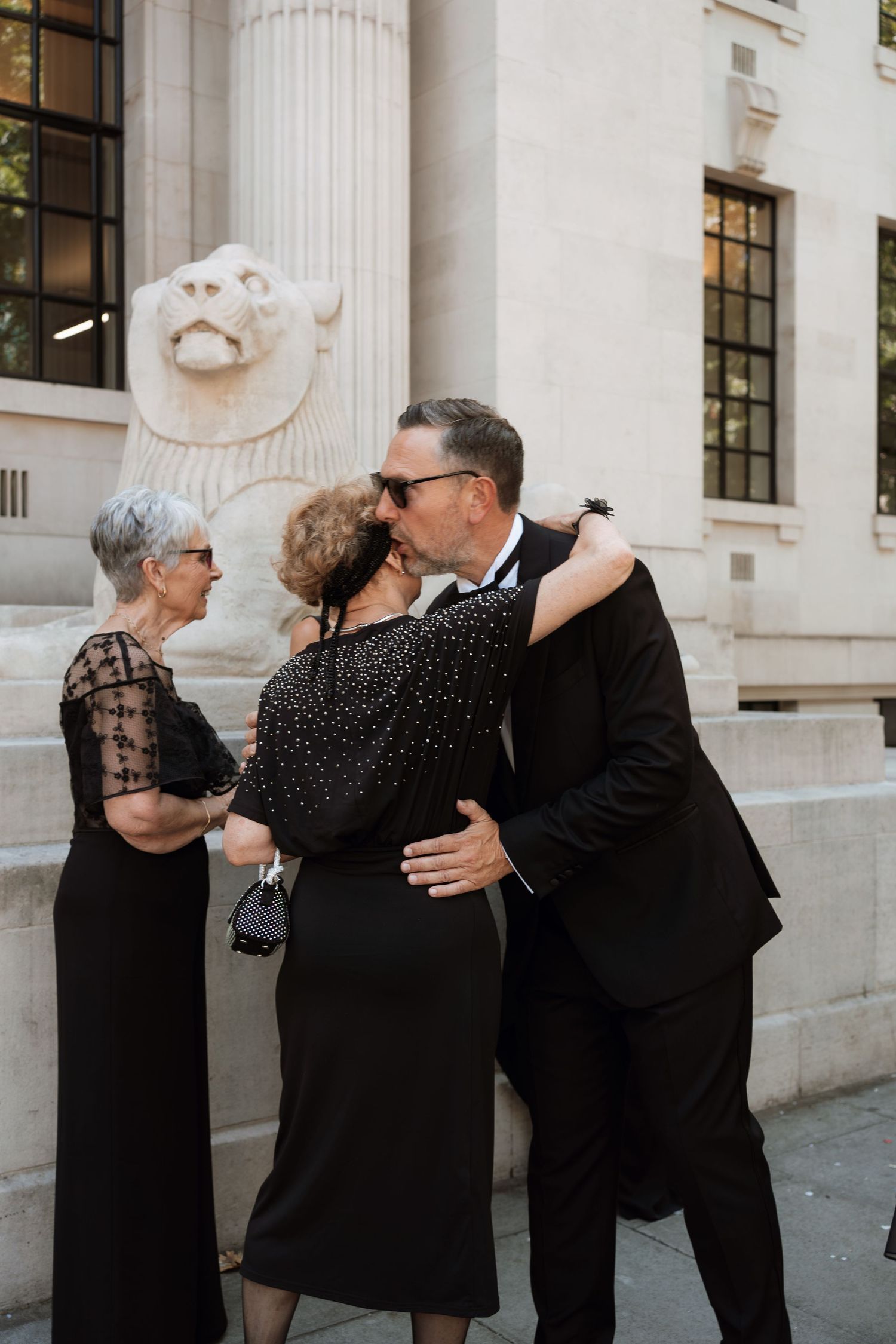 Three progressive photos show a couple in formal black attire embracing outside a classical building with columns.