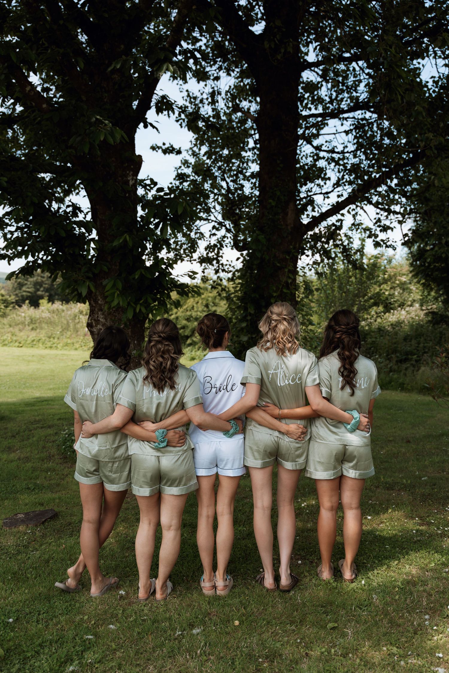 A group of bridesmaids in matching outfits standing with their backs to the camera on a grassy lawn.