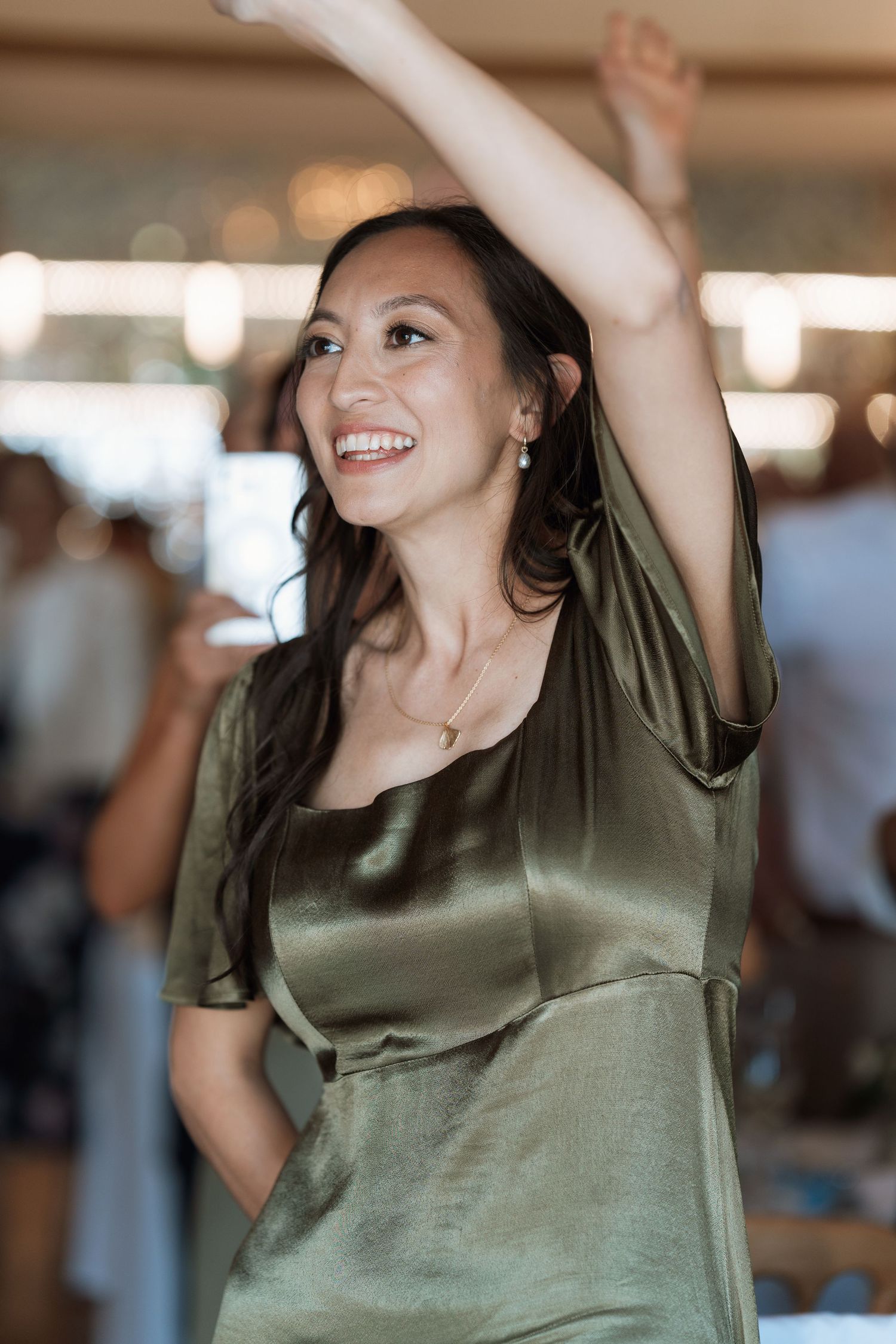 Guest in olive silk dress dances with arms raised at wedding reception celebration.