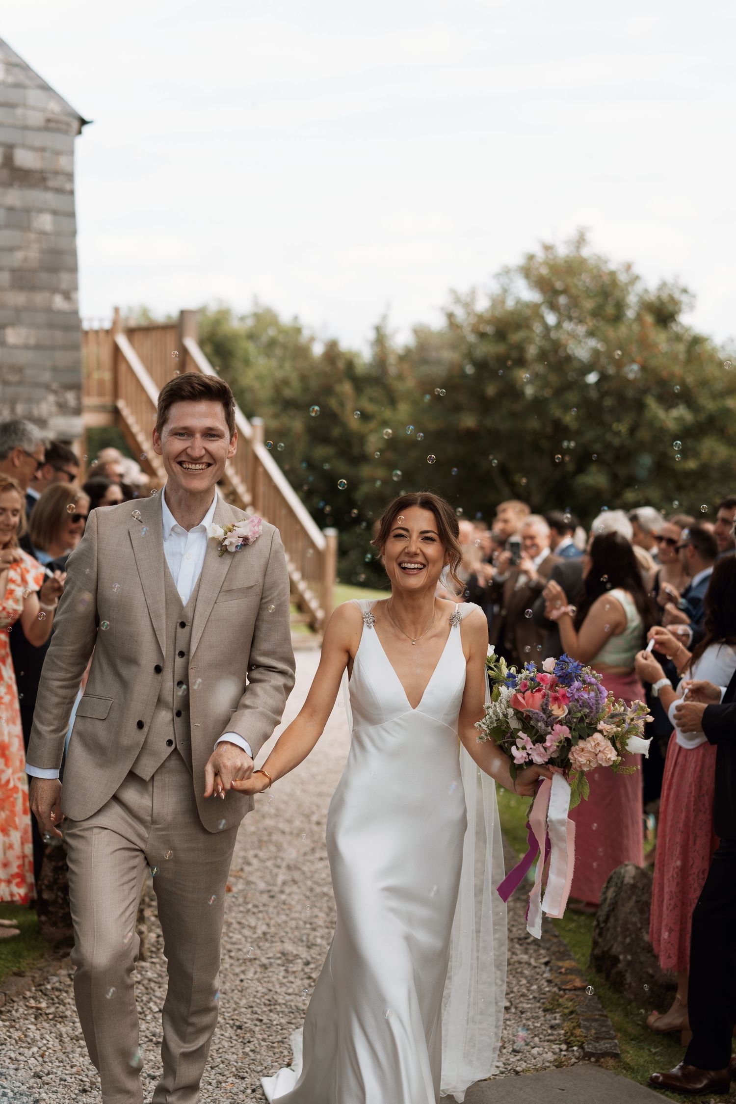 Newlyweds walking down the aisle while guests celebrate at an outdoor stone venue.