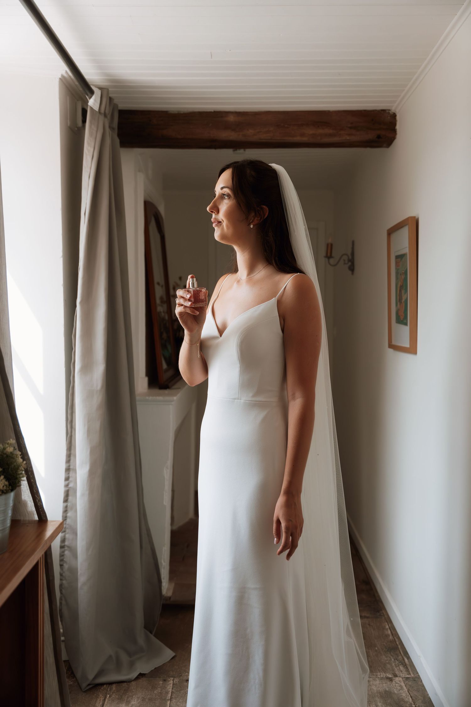 A bride in a classic white gown stands in a minimalist white hallway with natural lighting.