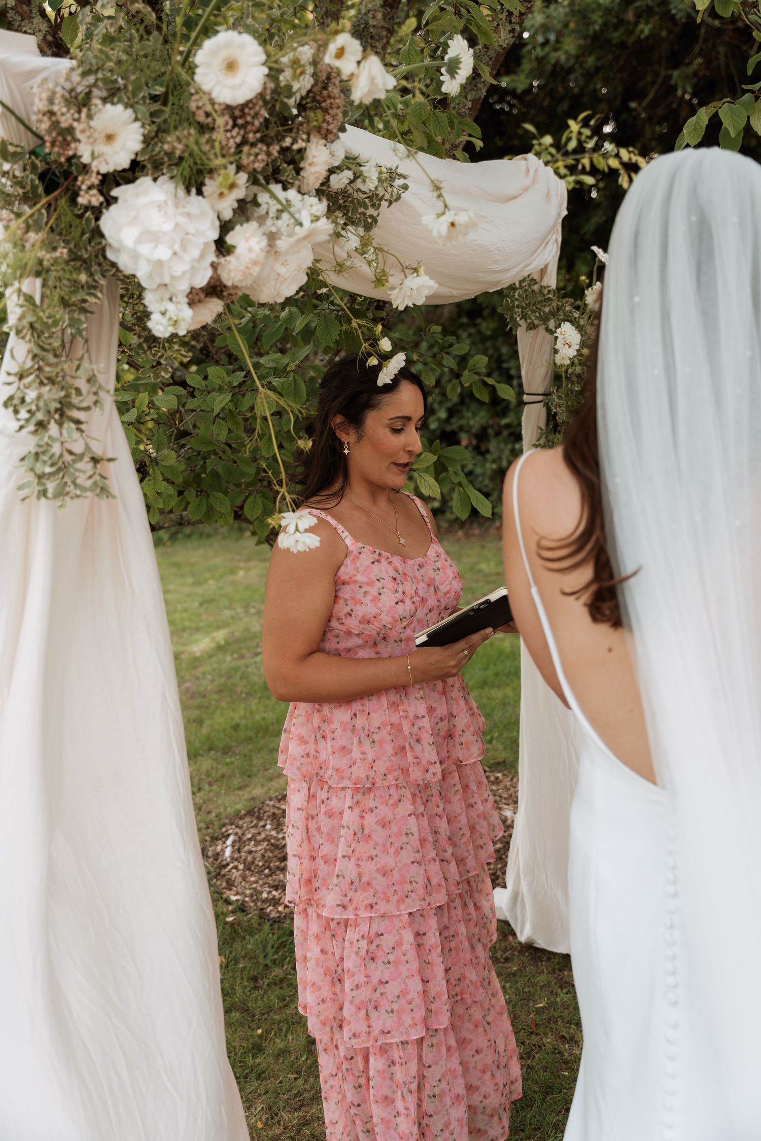 Wedding party members in pink dresses stand under white fabric and floral decorations.