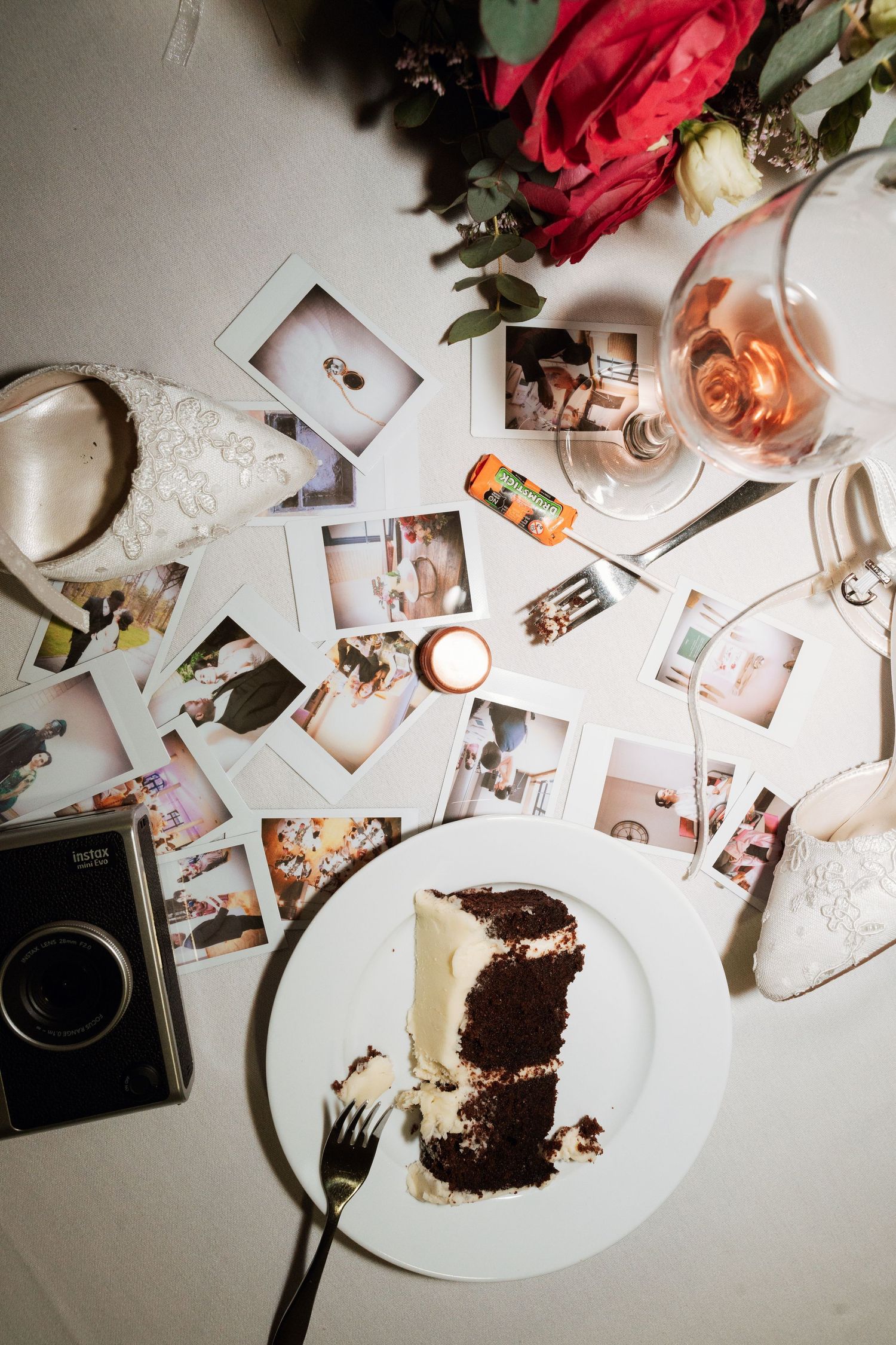 Overhead view of Instax photos, chocolate cake slice, rose wine, and red flowers on a white table.