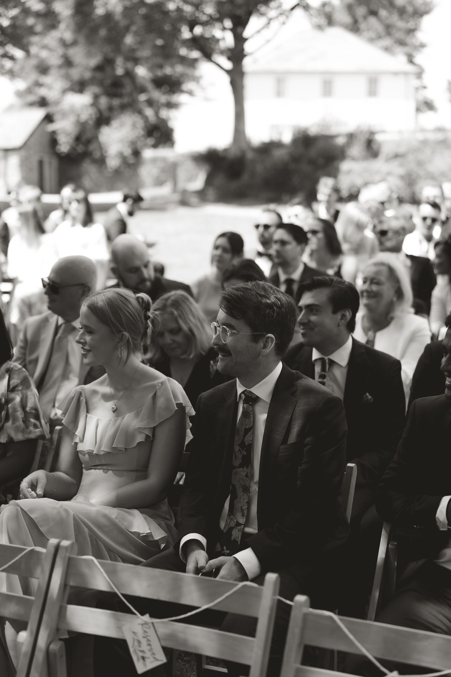 Wedding ceremony guests seated in rows of wooden chairs outdoors.