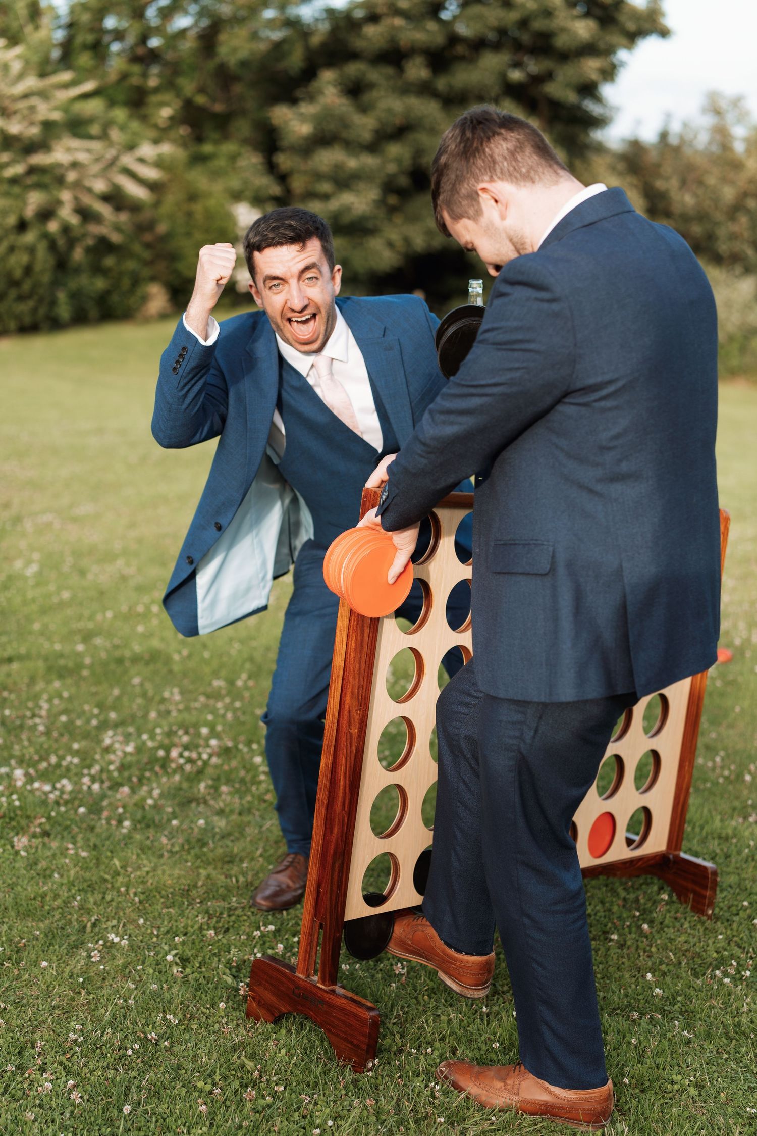 People in suits playing giant wooden connect four game on grass.