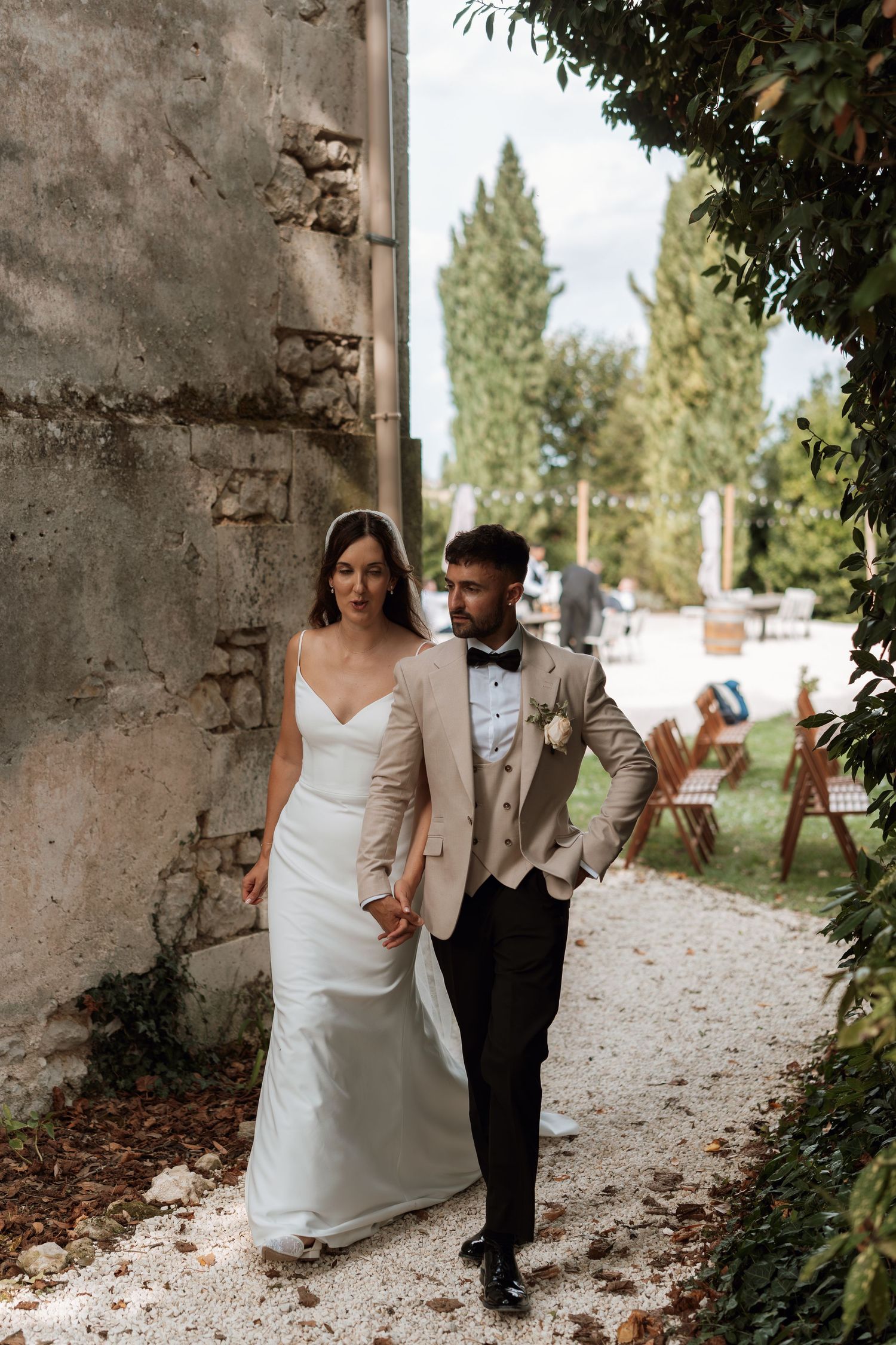 A couple walks together along a stone path bordered by rustic walls and greenery.