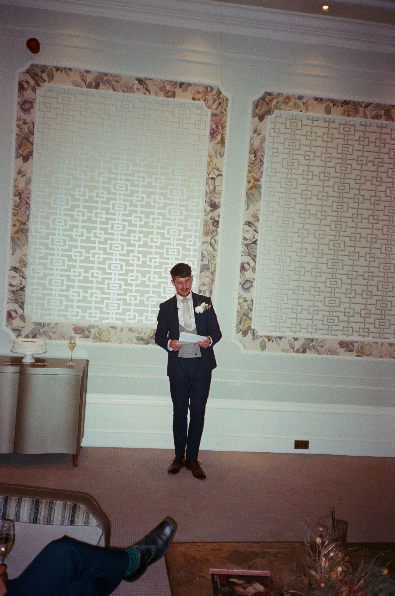 A person in formal attire stands at a podium in a banquet hall with patterned wallpaper.