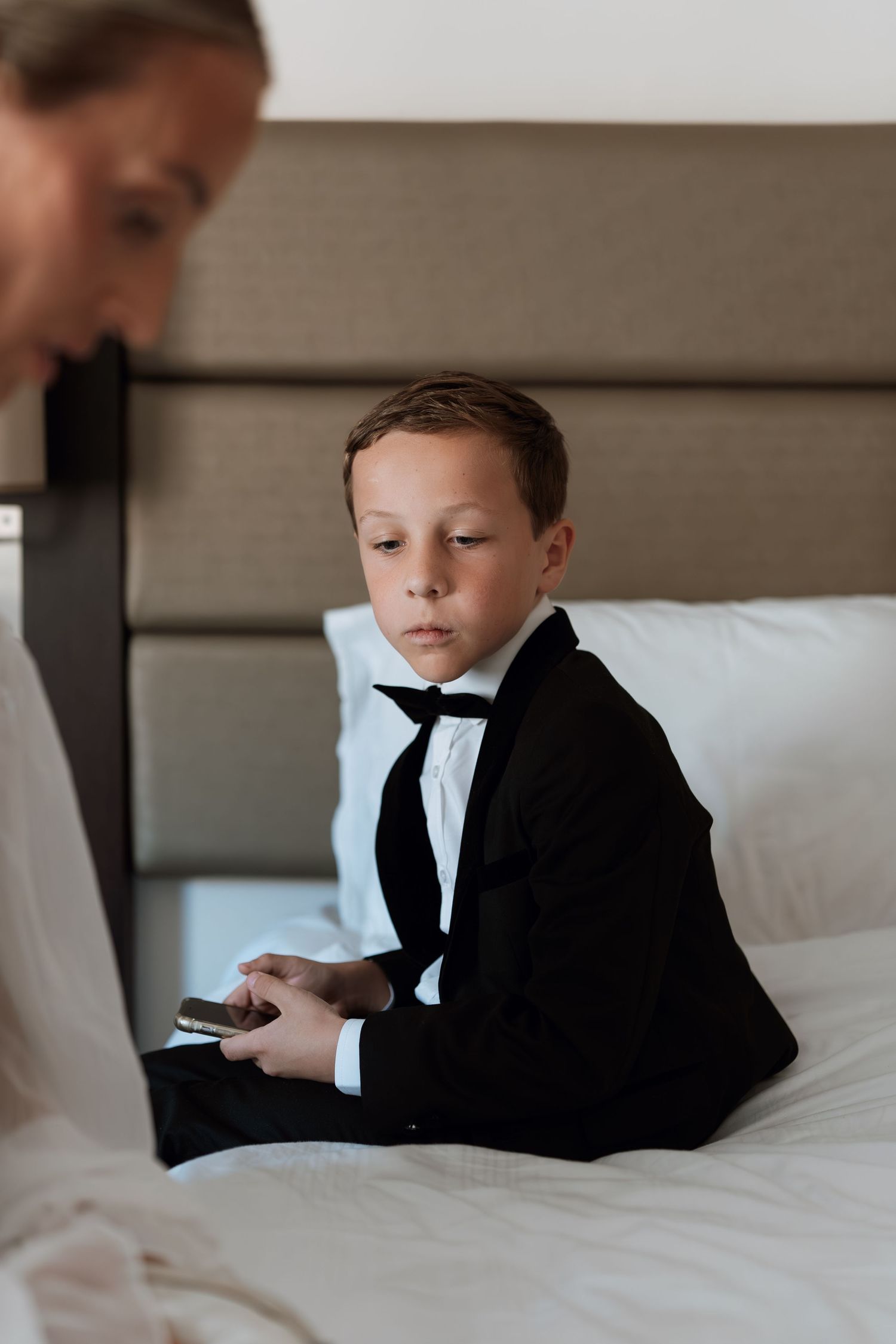 A young boy in a formal black suit and bowtie sitting on a white bed.