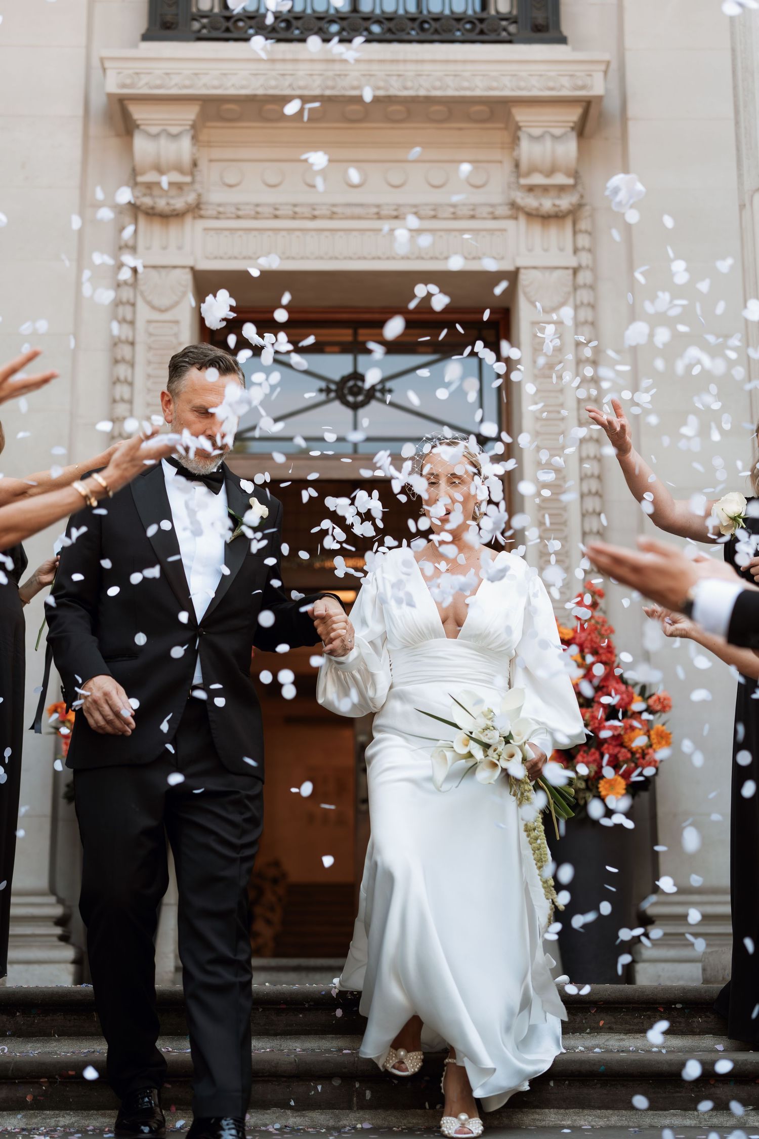 Newlyweds exiting building amid shower of white confetti petals.