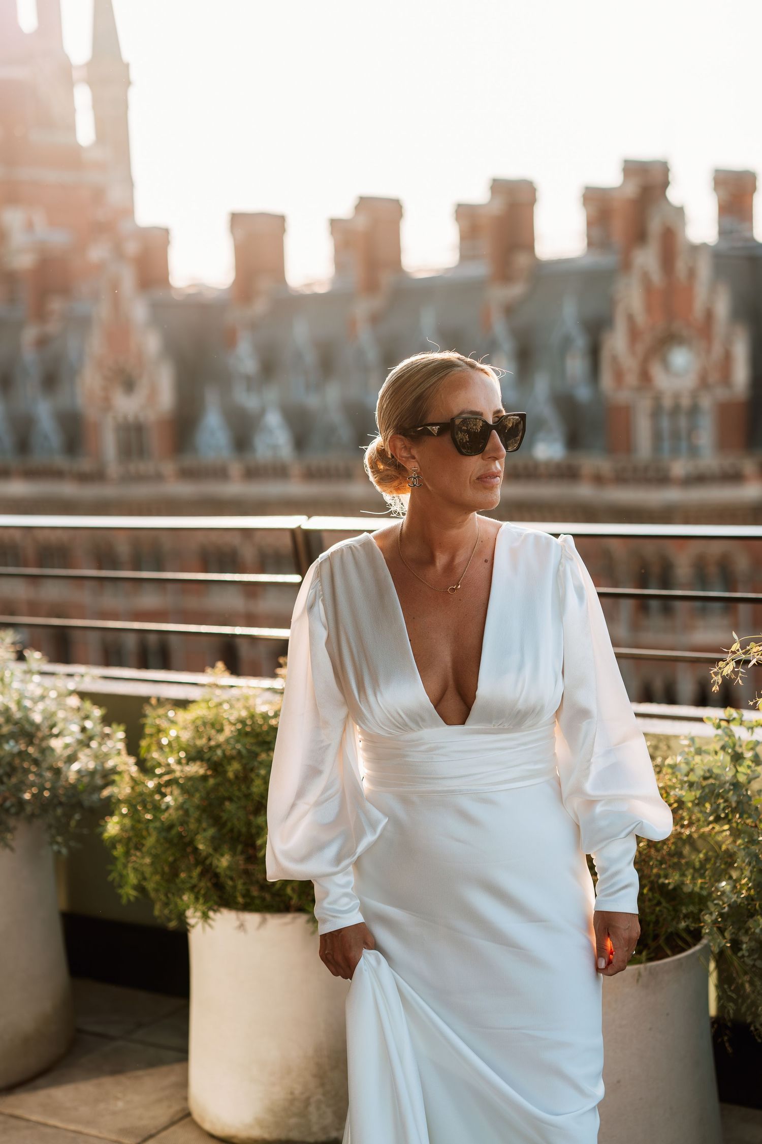 A person in a white dress poses on a rooftop terrace at sunset with city buildings in background.