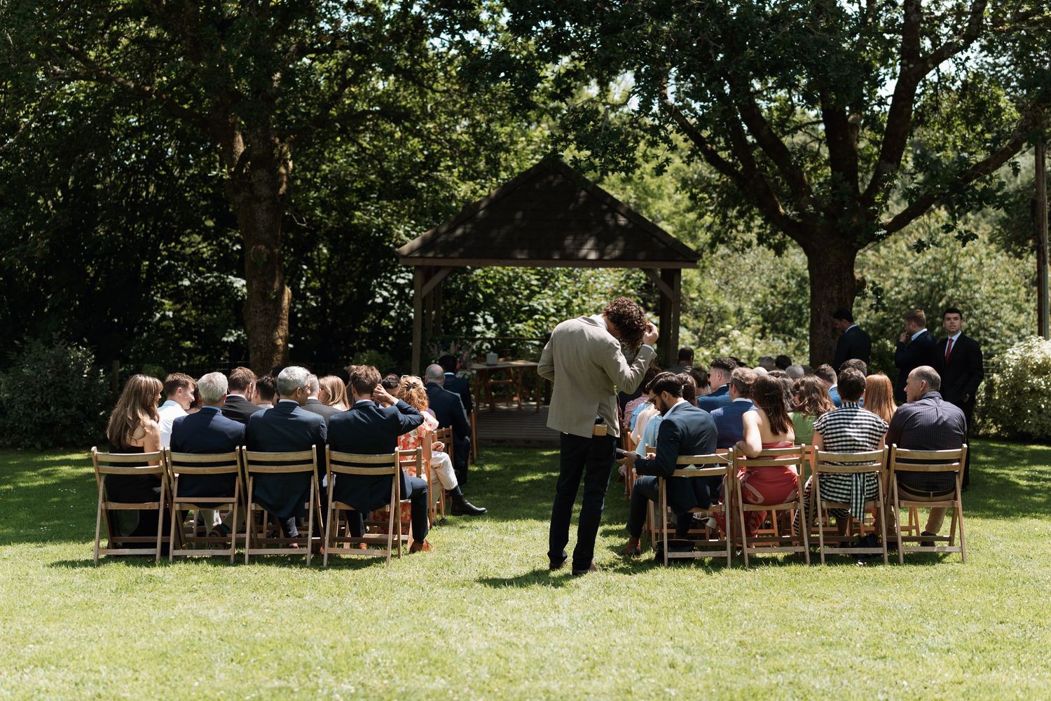 Wedding guests seated in wooden chairs on a sunny lawn surrounded by trees.
