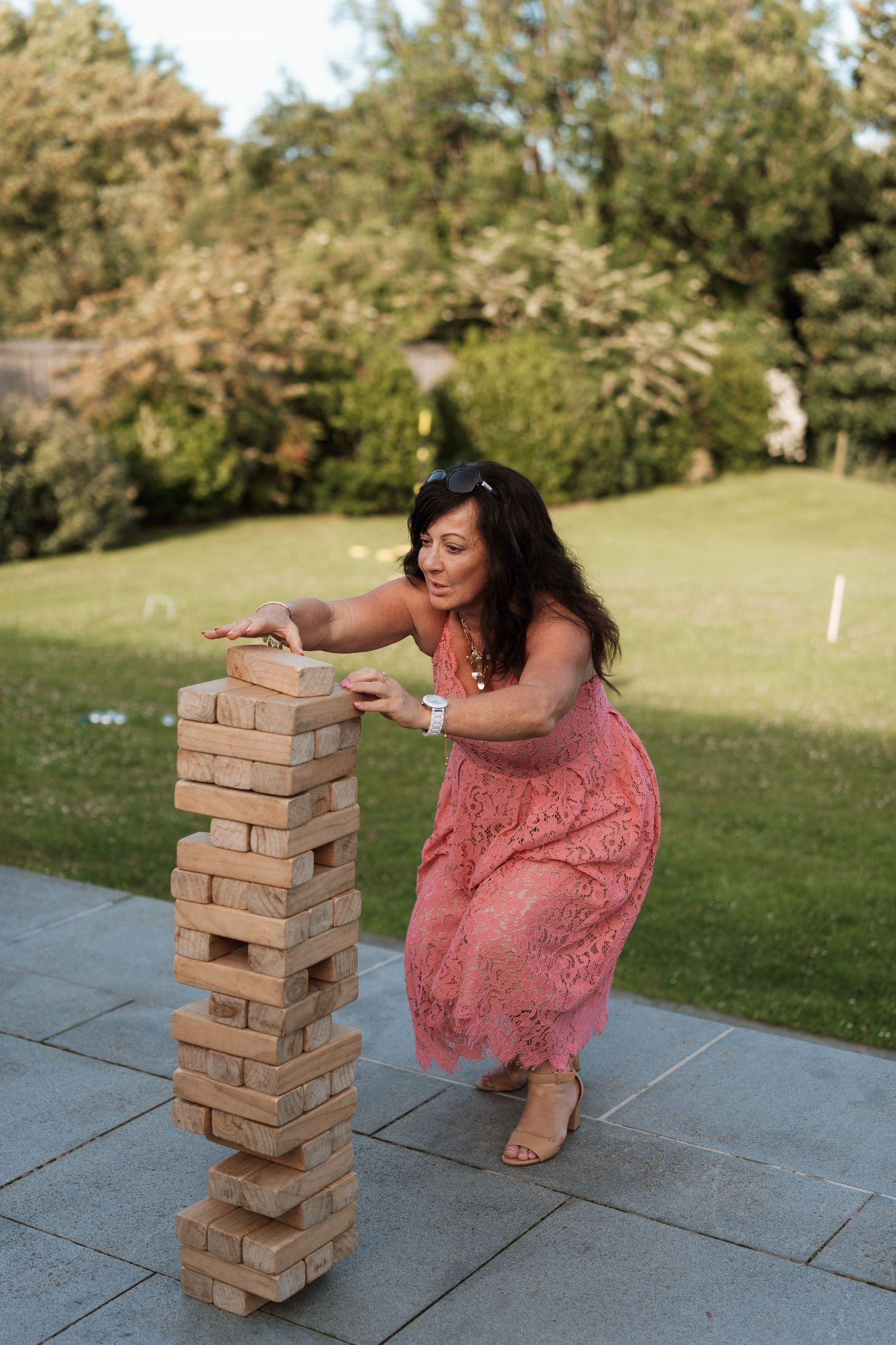 A person in a coral dress plays a game of giant Jenga on an outdoor patio during a sunny day.