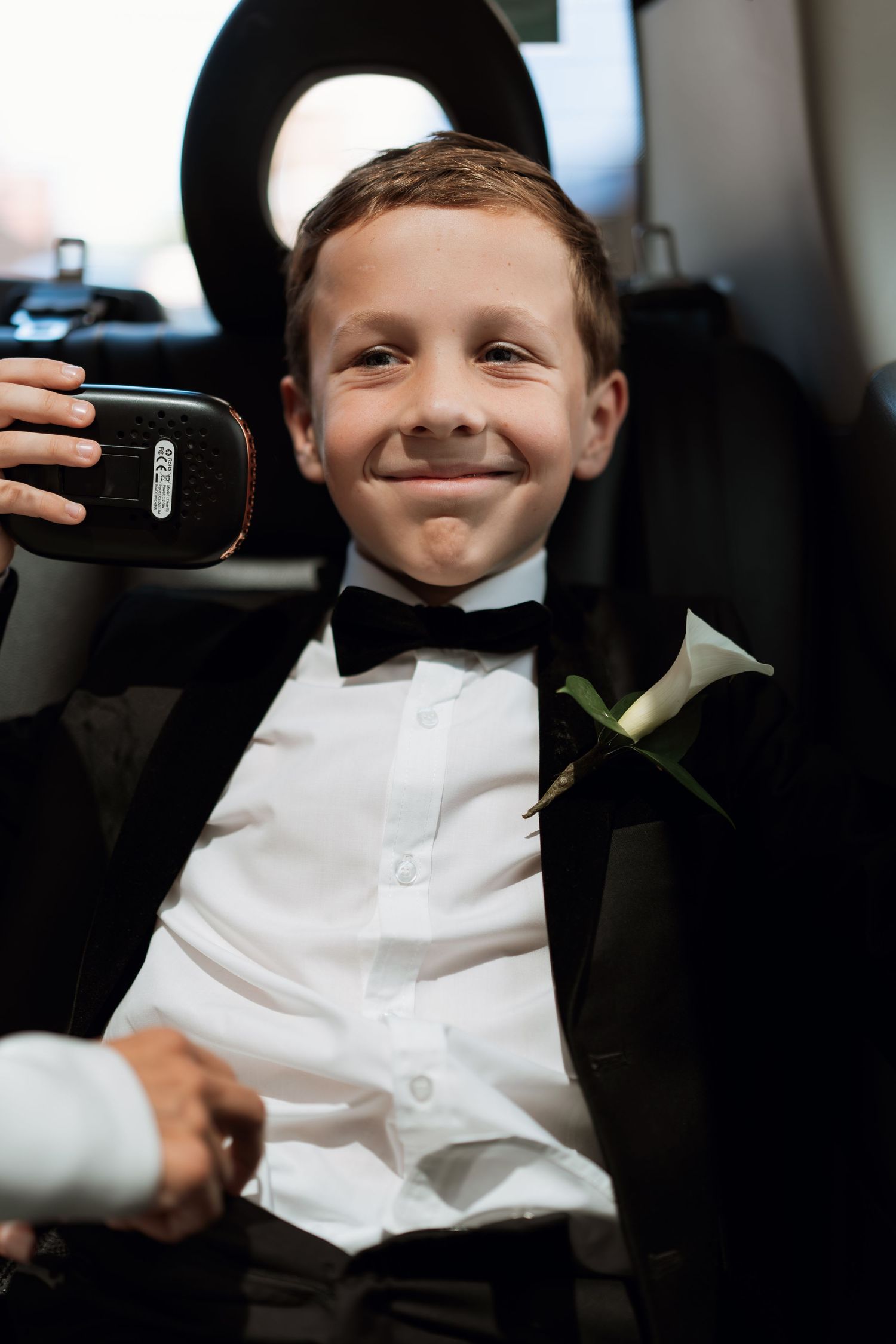 Young ring bearer in formal white attire sitting in a car during a wedding.