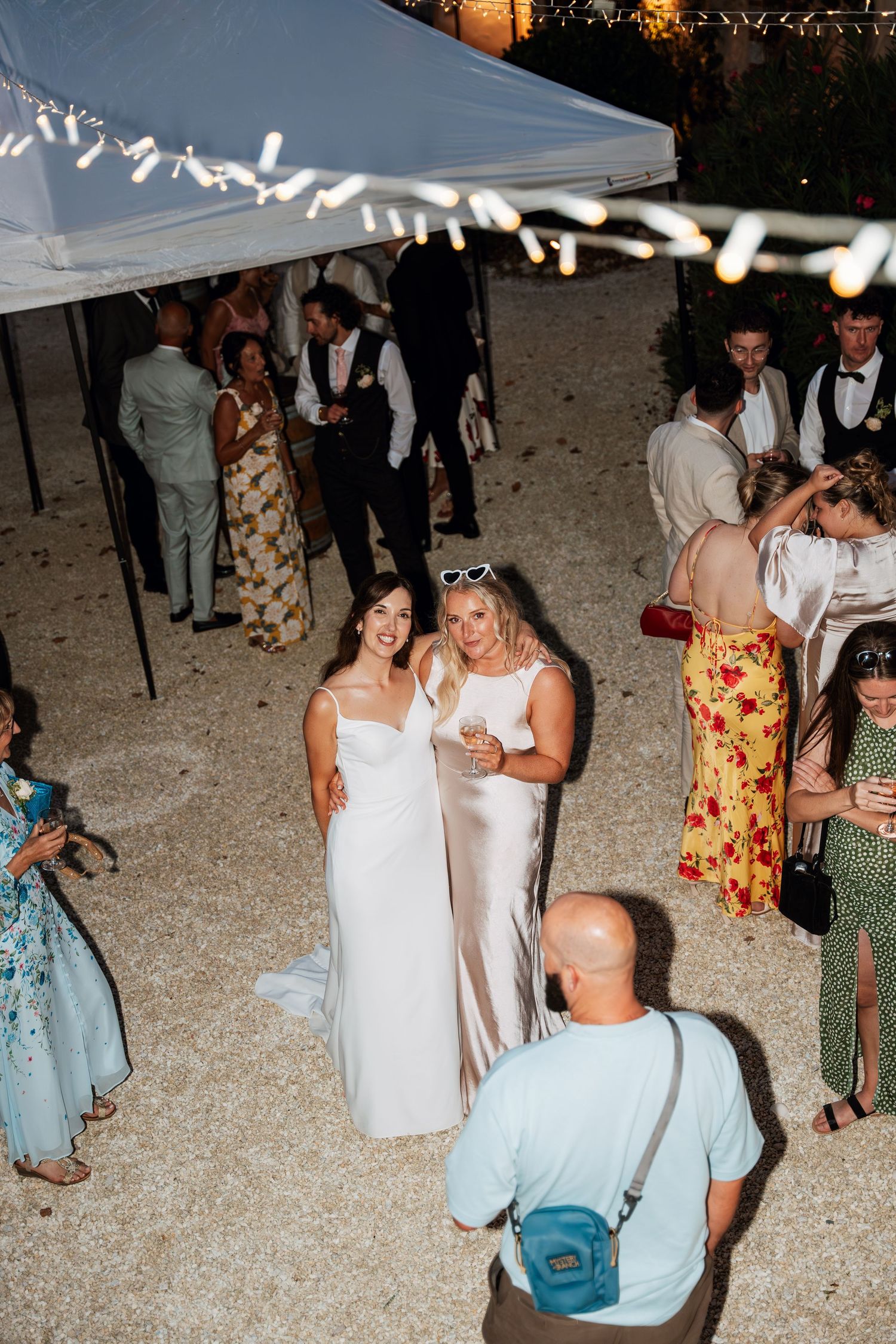 Wedding guests dancing and celebrating on a beach at sunset under string lights and a white tent.