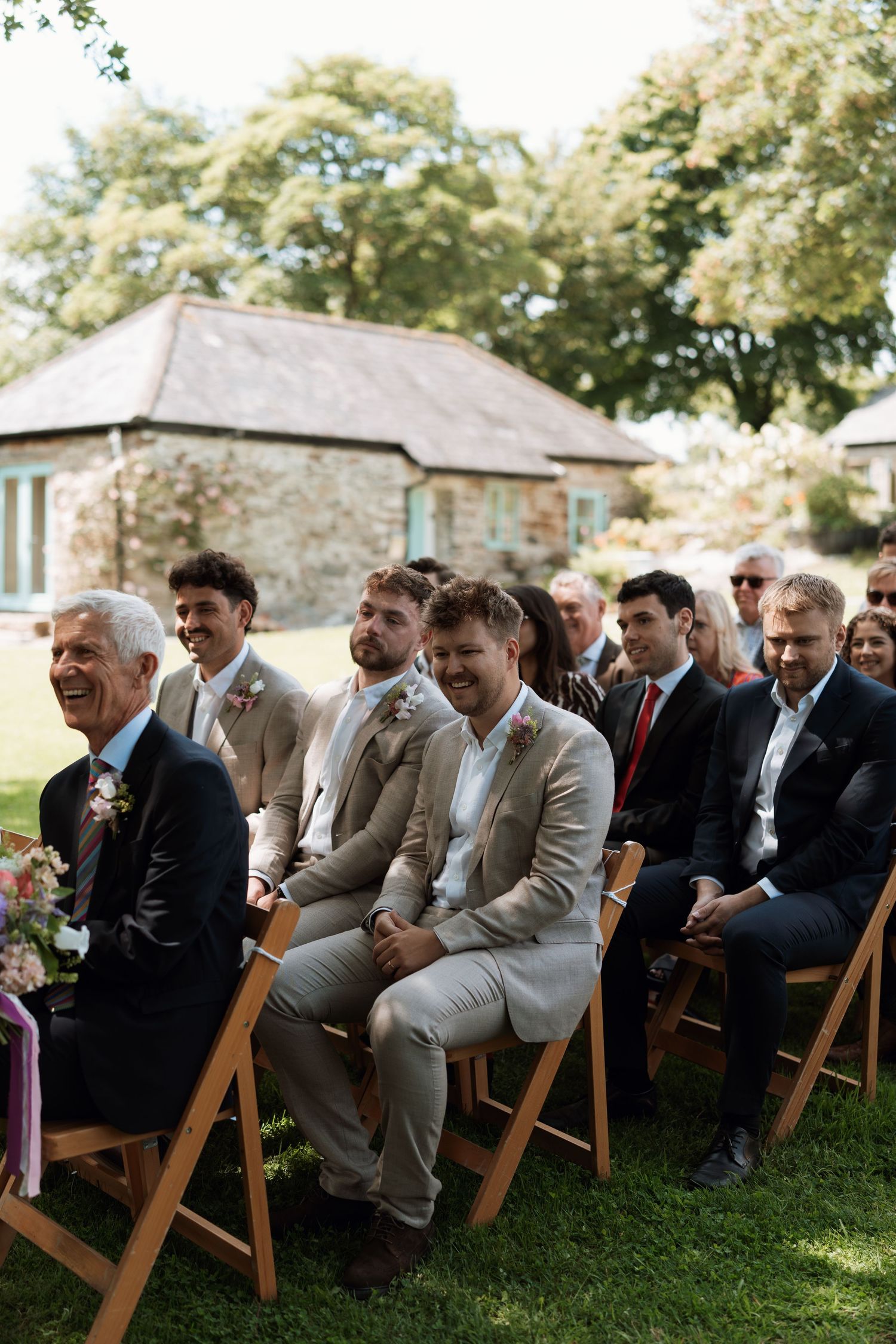 Wedding guests in formal attire seated on wooden chairs during an outdoor ceremony.
