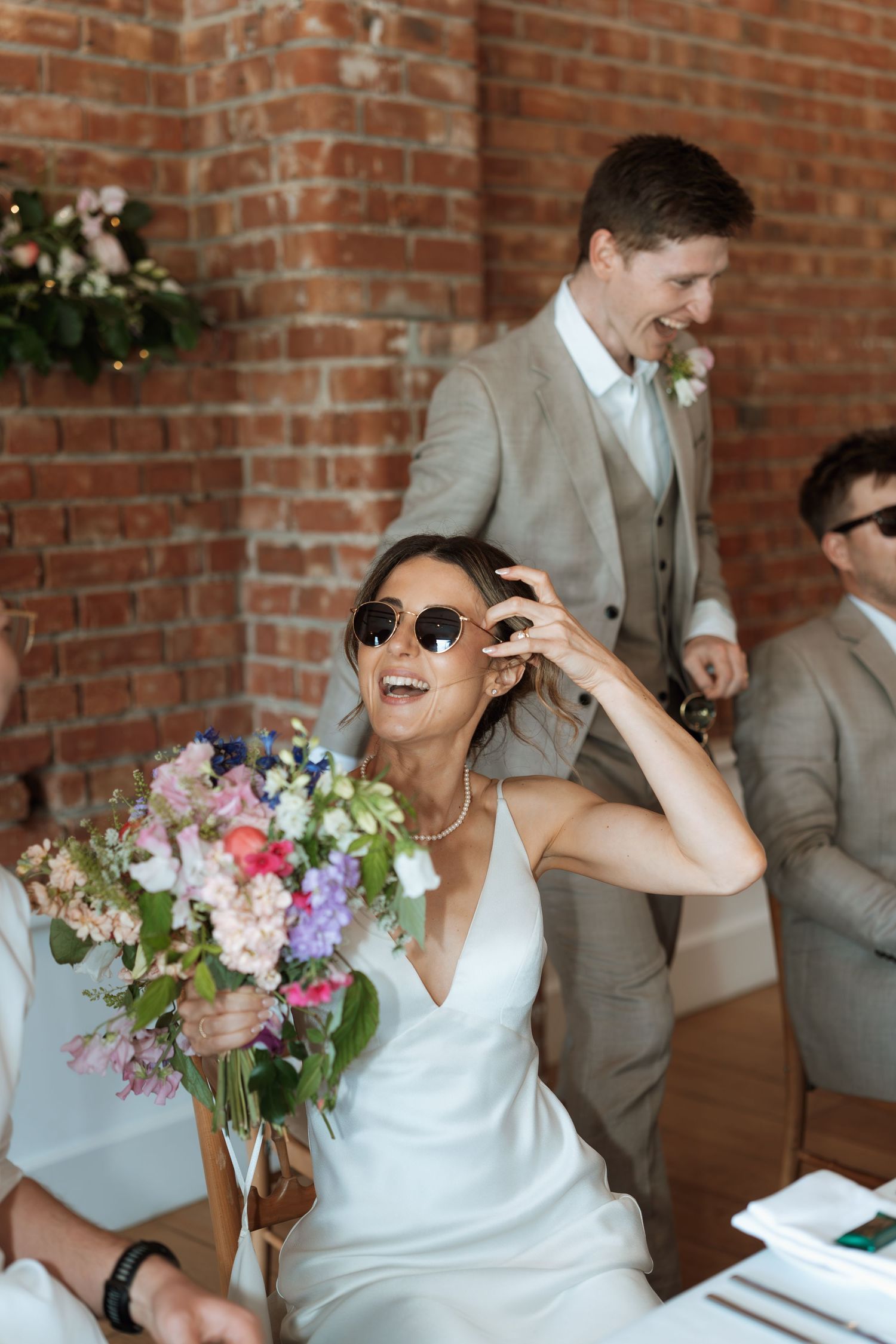 Wedding party members laughing while holding bouquets against brick wall backdrop.